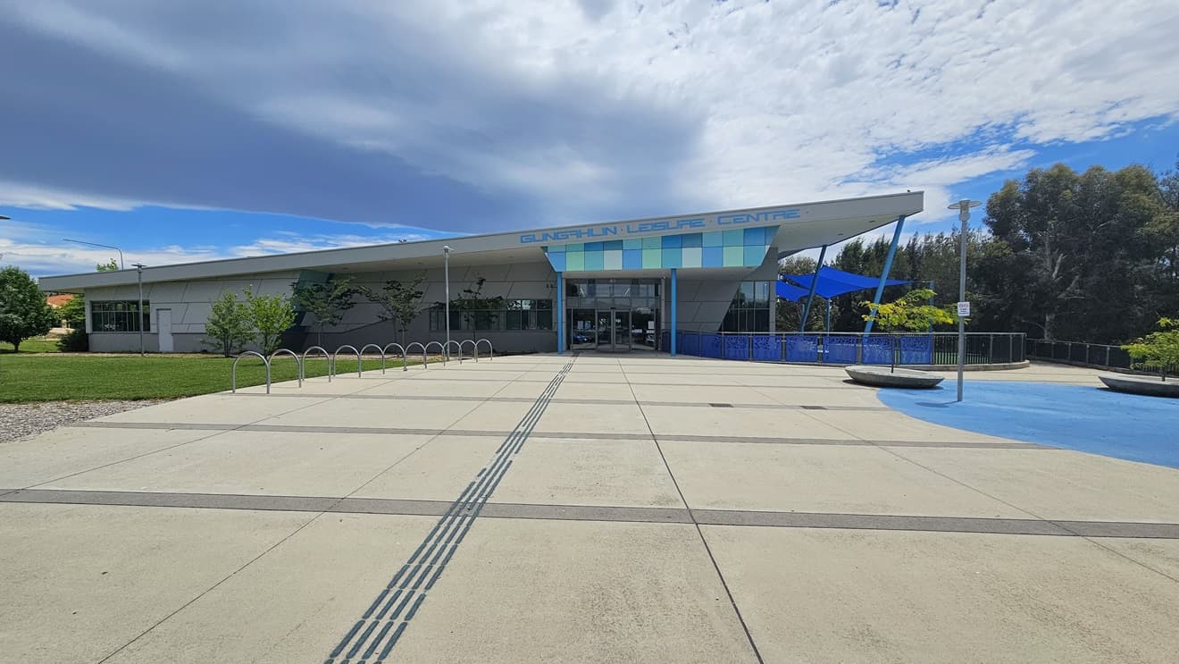 The building is a low-rise concrete structure with contrasting blue trim, and the facility name is displayed at the entrance. The building's concourse features a spacious concrete area. On the left, bike racks are positioned near the grass area. To the right, two small trees are in large concrete pots. The concrete floor has blue markings. The building has a blue fence railing and blue shade cloths. Tall trees fill the area on the right.