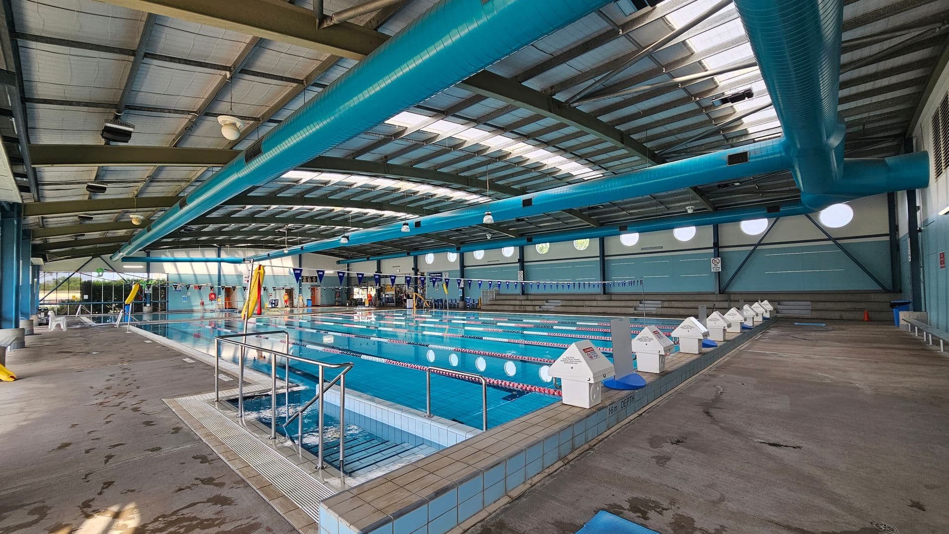 An indoor swimming pool with multiple lanes, marked by red and white lane dividers. On the right, a yellow slide is visible near the pool's edge. The pool is surrounded by a wet concrete floor with depth markers. On the left, there are blue chairs and a wall with circular windows and signs. The ceiling is high with exposed beams and large blue ventilation ducts. The far end of the pool has a lifeguard chair and more seating.
