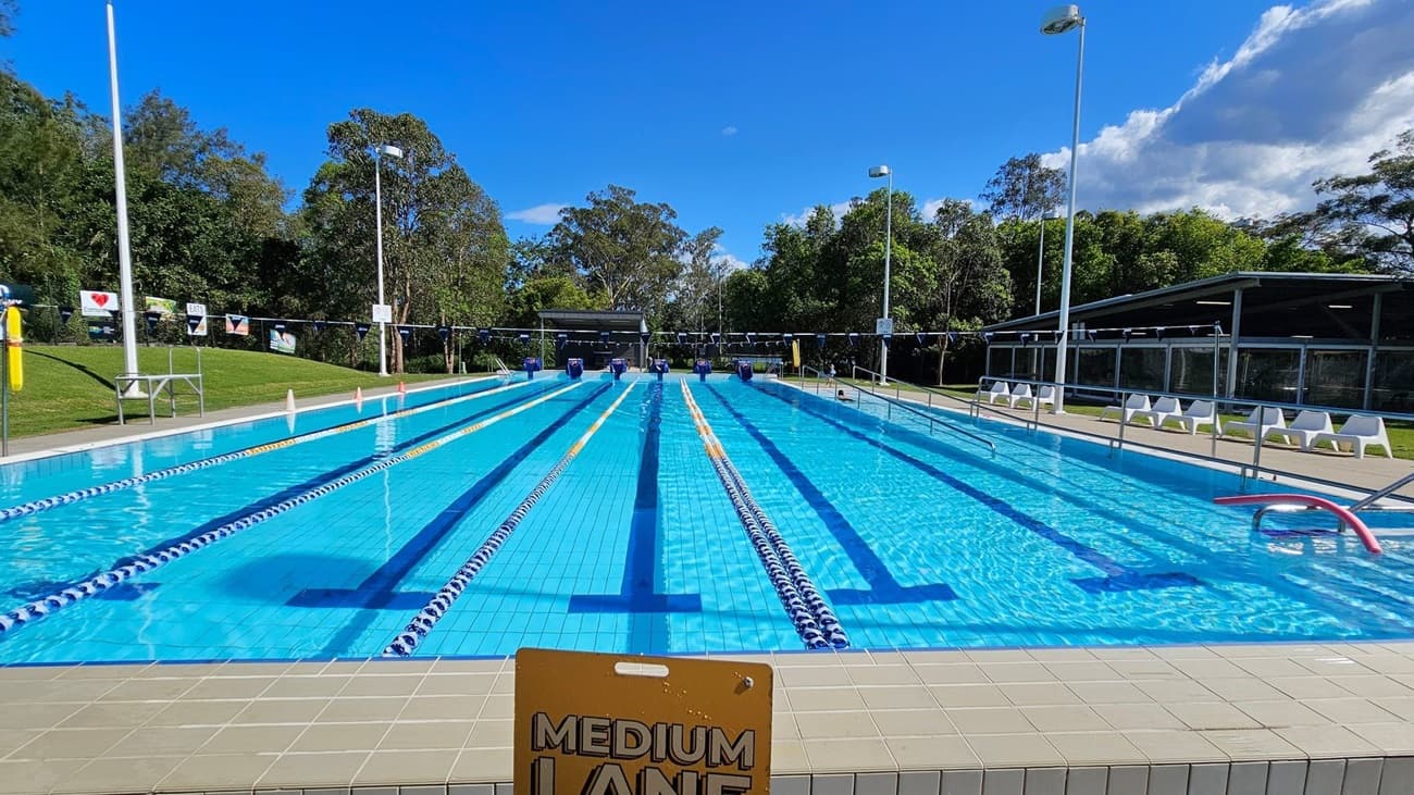 An outdoor swimming pool with multiple lanes separated by lane dividers. In the foreground, a sign reads "MEDIUM LANE." The pool is surrounded by a tiled deck. On the left, there is a grassy area with a fence displaying various signs. On the right, there are white plastic lounge chairs and a covered structure. In the background, there are trees and tall light poles.