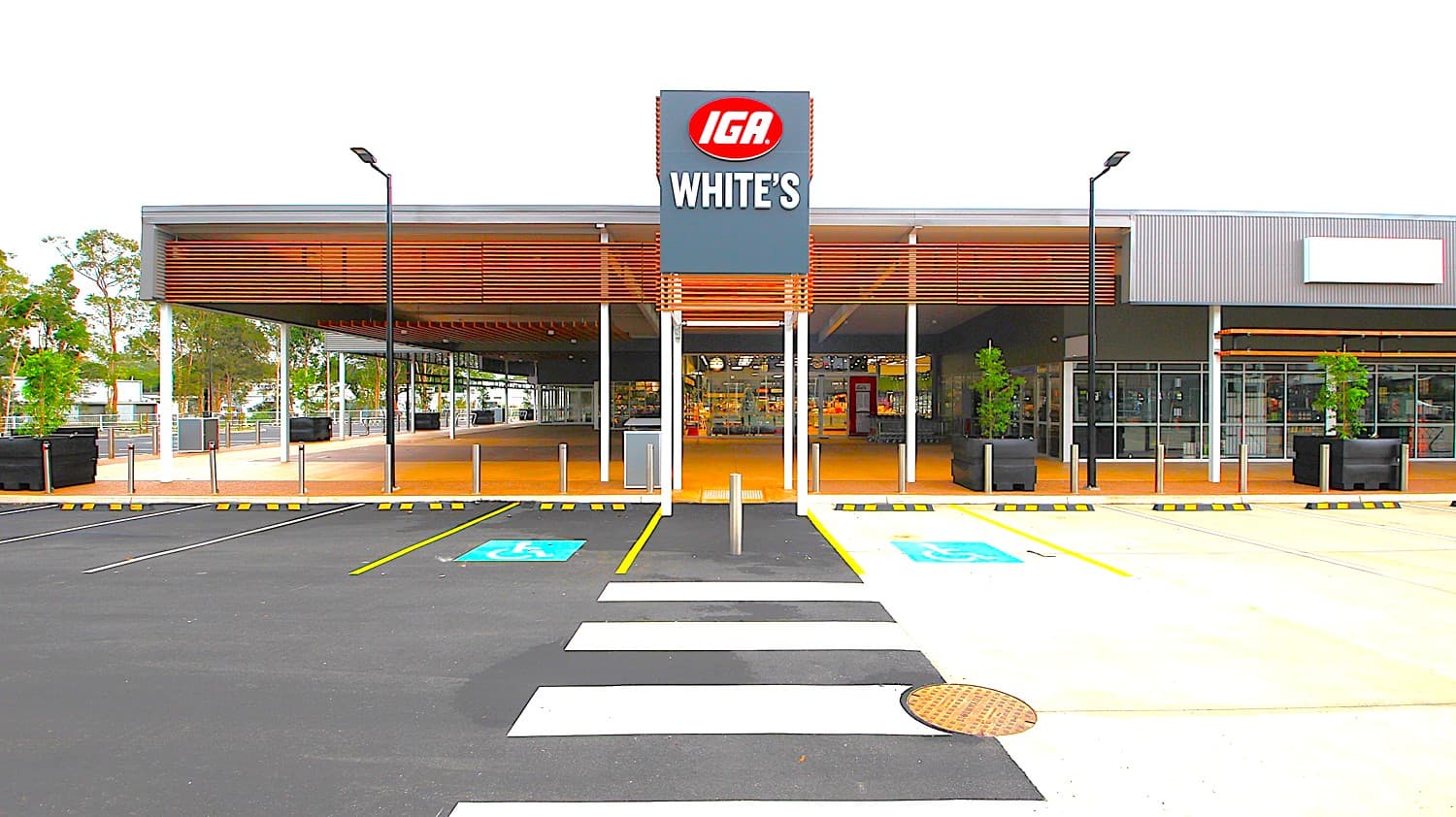 A grey-painted store building with a wide awning on the left and glass designed store on the right. A promenade with short stainless steel pillars overlooks the large parking lot, which is delineated by a pedestrian-marked path with a White's IGA Forest Glen billboard. Big pots plants are seen round and trees outside creating a quiet scene.