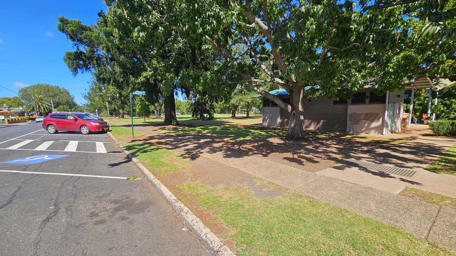 A red car is parked on the left side near a zebra crossing on an asphalt road. A blue disabled parking sign is visible on the road. On the right, there is a building with a grey wall and a corrugated metal roof, partially obscured by a large tree with dense green foliage. The ground is a mix of grass and paved pathways. In the background, more trees and a grassy area are visible, with a clear blue sky above.