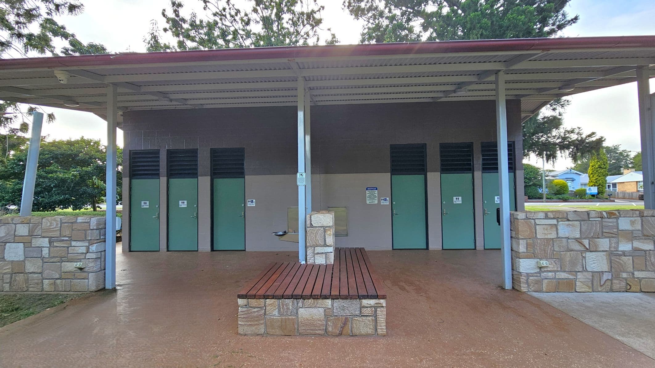 A public restroom facility with a metal roof and stone walls. There are three green doors with ventilation panels above them, each marked with restroom signs. In the centre, a stone and wood bench is positioned in front of a supporting pillar. A drinking fountain is attached to the pillar. The ground is paved, and trees and a few buildings are visible in the background.