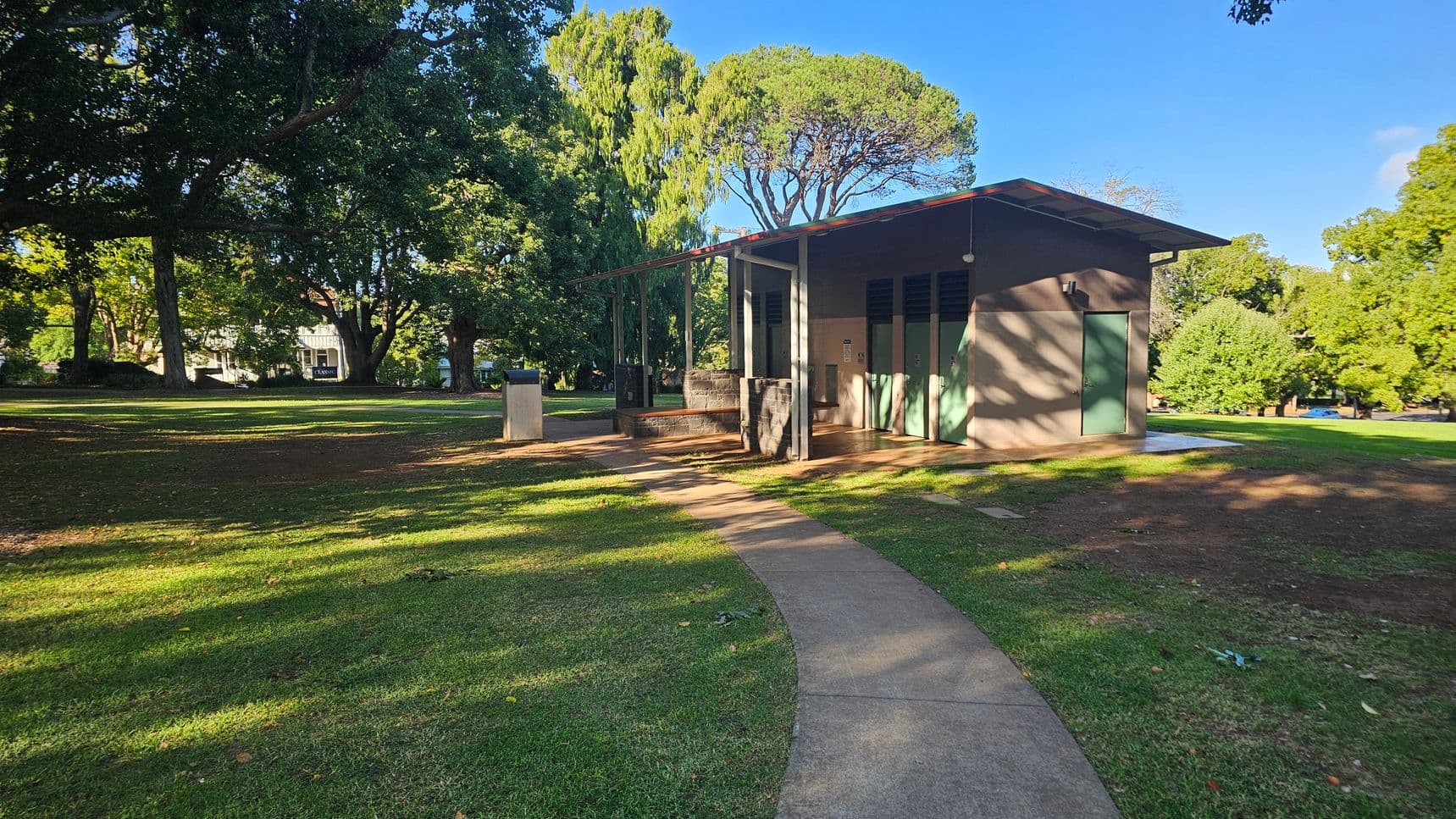 A park scene with a small building on the right, featuring green doors and a sloped roof. A curved concrete path leads to the building, surrounded by grass. Large trees with dense foliage are in the background, casting shadows on the ground. A few structures, possibly benches or bins, are near the building. The area is well-lit, suggesting daylight.