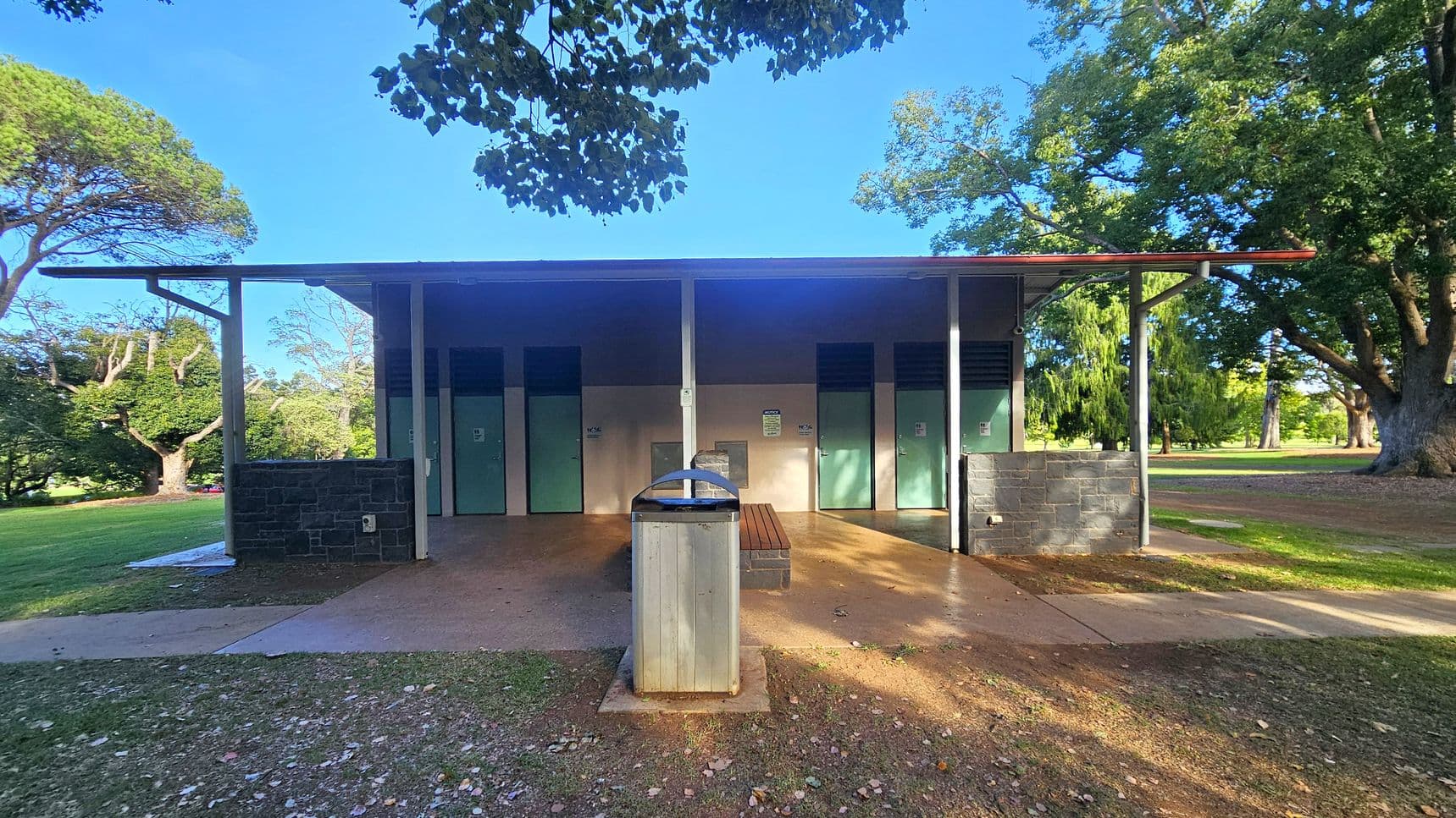 A public restroom building with a flat roof and green doors is in the centre. A rubbish bin is positioned in front of the building, with a bench to its right. The structure is flanked by stone walls on either side. The ground is a mix of grass and dirt, with scattered leaves. Large trees with dense foliage surround the area, providing shade. The sky is clear and blue.