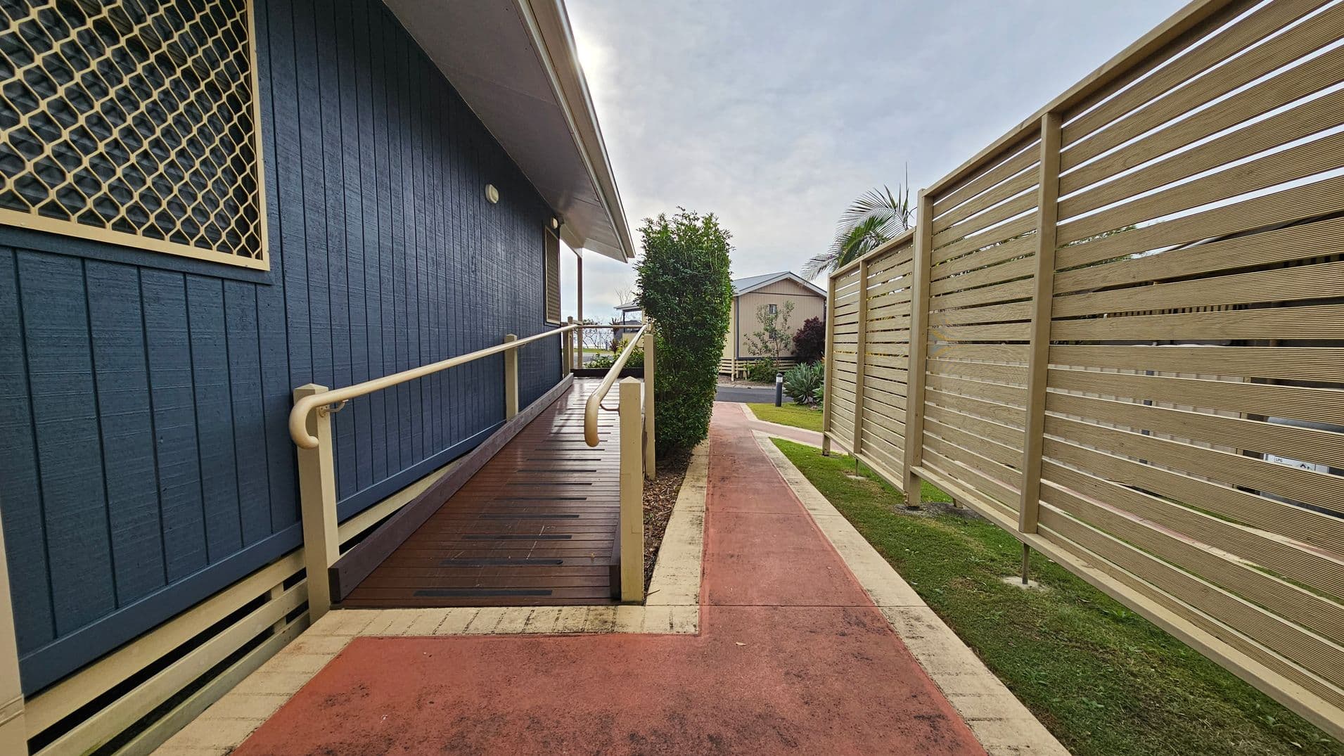 A pathway with a reddish surface runs between a blue building on the left and a beige slatted fence on the right. The building has a ramp with a handrail and a window with a mesh cover. A small bush is visible at the end of the path, with another building in the background. The ground is grassy alongside the path.