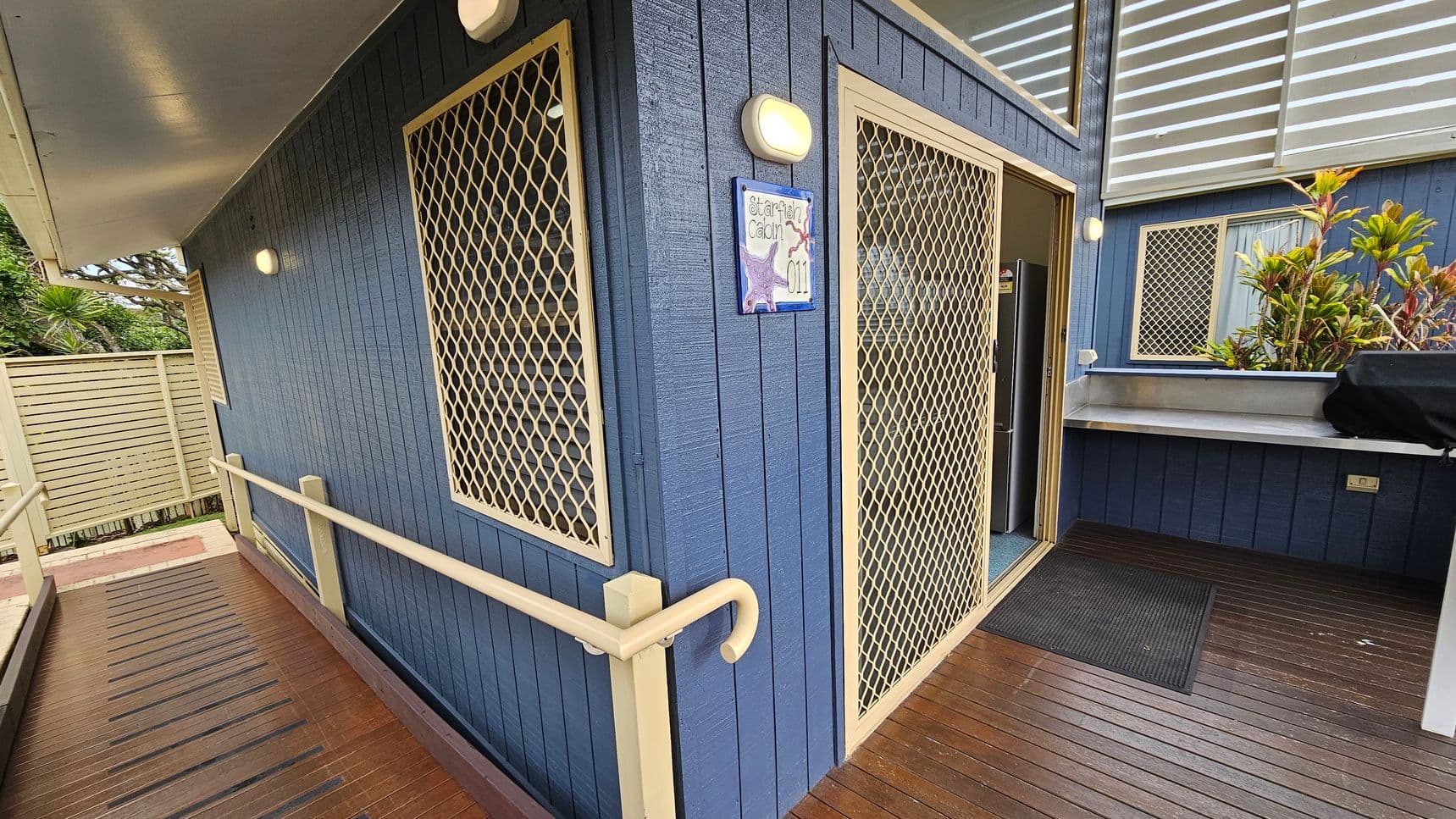 A blue cabin with cream-coloured mesh doors and windows. A sign on the wall reads "Starfish Cabin 011" with starfish decorations. A wooden ramp with a cream handrail leads to the entrance. To the right, there's a metal countertop with a black cover, possibly for a grill. Plants are visible near the window. The ceiling is light-coloured, and the floor is made of wooden planks.