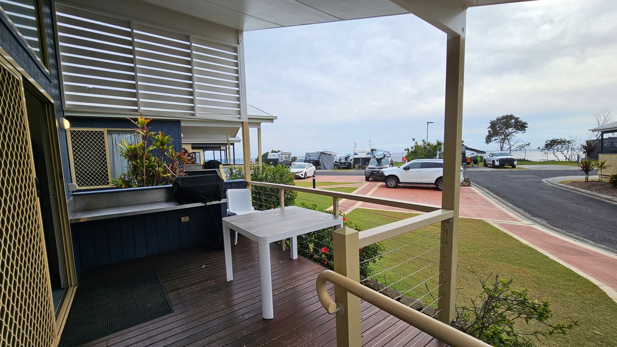 A wooden deck with a white table and chair is in the foreground, bordered by a railing. To the left, there's a barbecue grill and a small garden with plants. The background shows a parking area with several cars and caravans, and a grassy area with a few trees. The sky is overcast.
