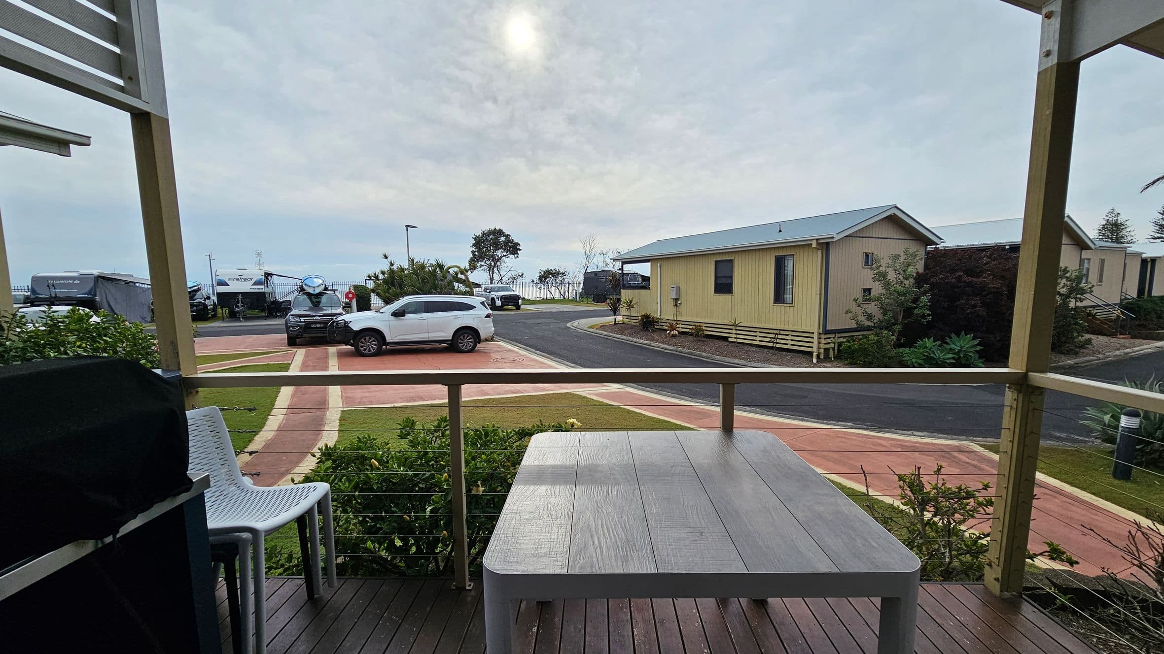 A wooden deck with a grey table and two white chairs in the foreground. To the left, a barbecue grill is partially visible. The background features a paved path leading to a road, with a white car parked nearby. A yellow cabin is on the right, surrounded by greenery. Further back, several caravans and a tree are visible, with the sea and a cloudy sky in the distance.