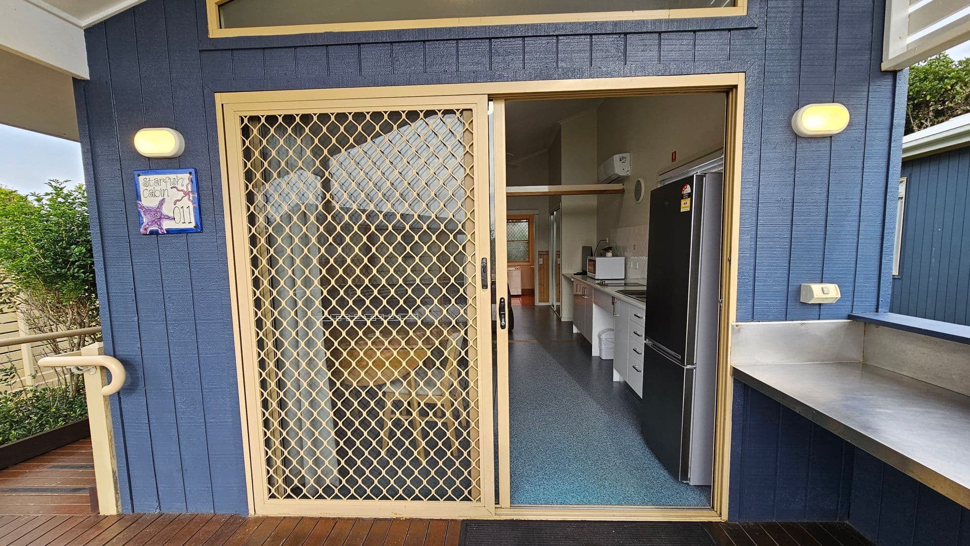 A blue exterior wall with a sliding mesh door leading into a kitchen. The door frame is light-coloured, and there's a small sign with a starfish design and the number "011" on the left. Inside, a black fridge and white cabinets are visible. The floor is a speckled blue, and a microwave sits on the counter. Outside, a wooden deck and a metal bench are present. Two wall-mounted lights are on either side of the door.