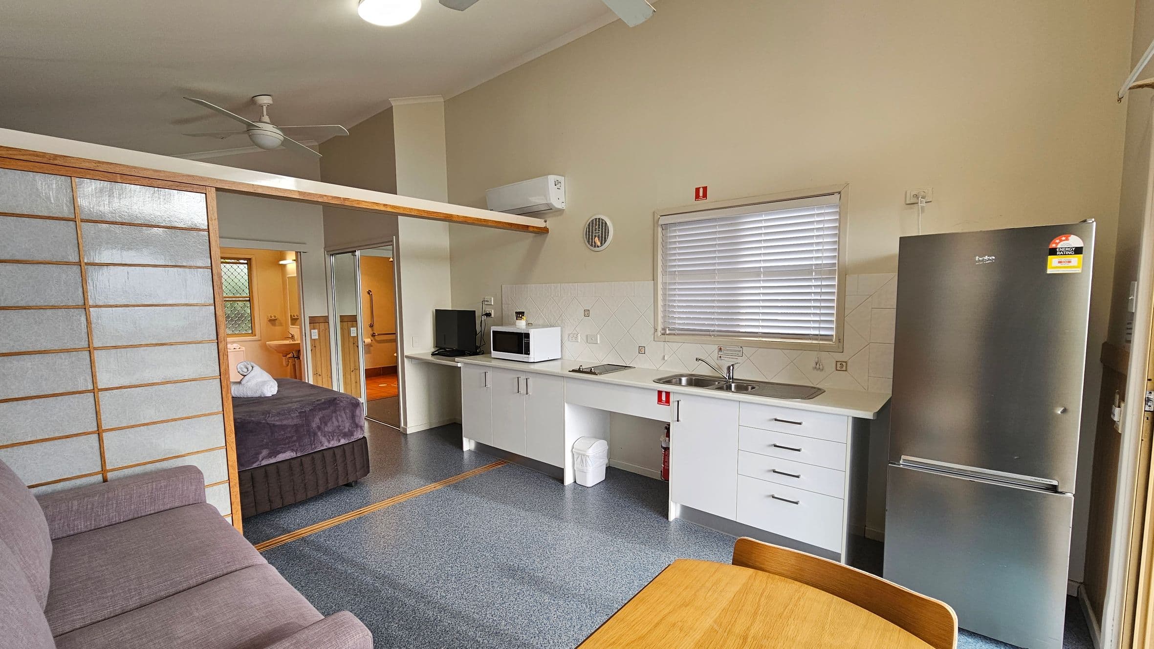 A compact living space with a kitchen on the right featuring white cabinets, a microwave, a sink, and a stainless steel fridge. A window with blinds is above the sink. In the foreground, a wooden table is partially visible. On the left, a purple sofa is next to a sliding partition that leads to a bedroom with a bed and a towel. The floor is dark grey, and the ceiling is light with a ceiling fan. A bathroom is visible through an open door in the background.
