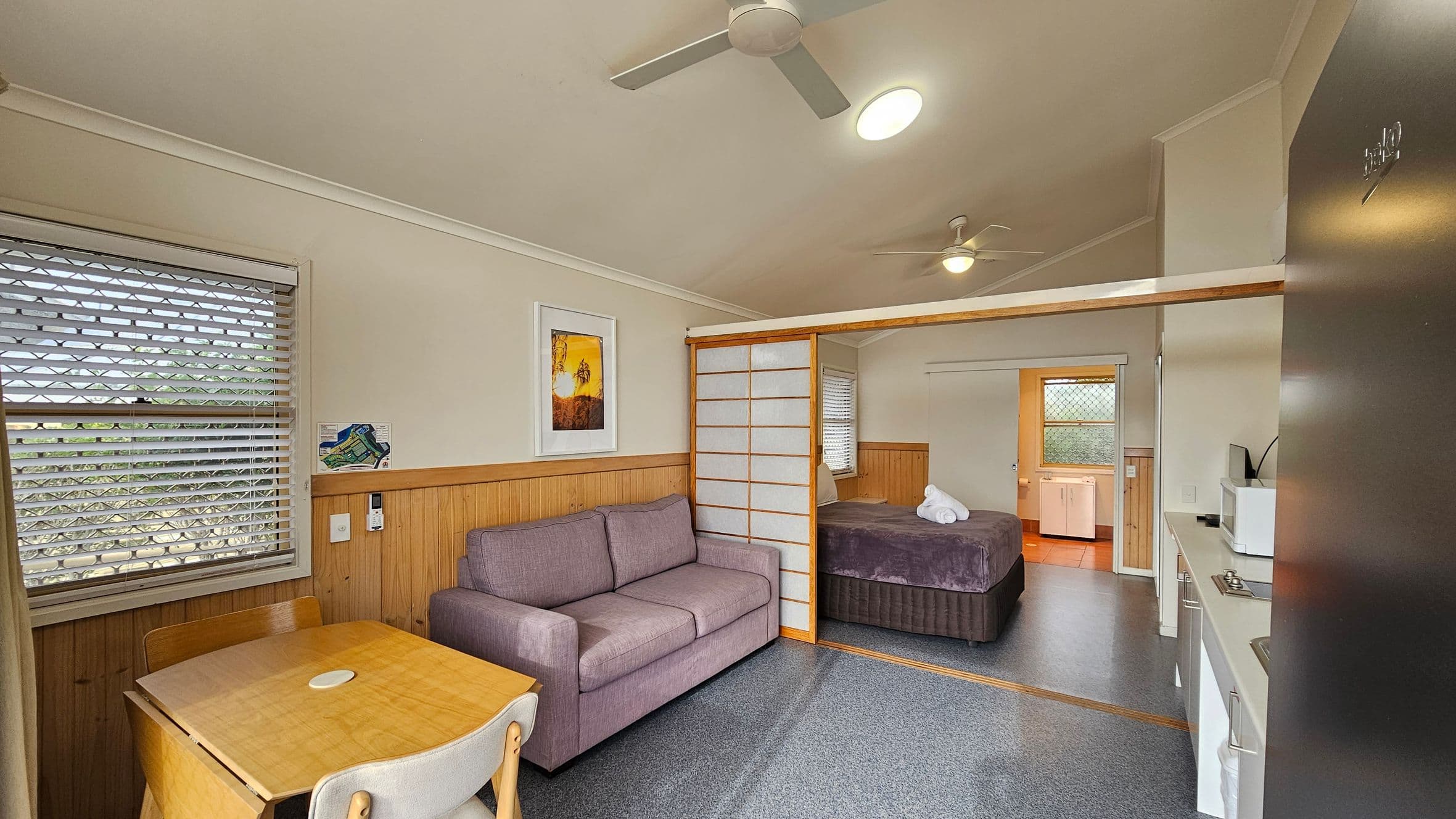 A compact room with a light purple sofa on the left, next to a small wooden table and two chairs. A window with blinds is above the table. A framed picture hangs on the wall above the sofa. In the centre, a sliding partition separates the living area from a bedroom with a bed covered in a dark blanket and neatly folded towels. On the right, a kitchenette with a microwave and sink. The ceiling has a fan and a light fixture. The floor is carpeted, except for the tiled area near the bathroom.