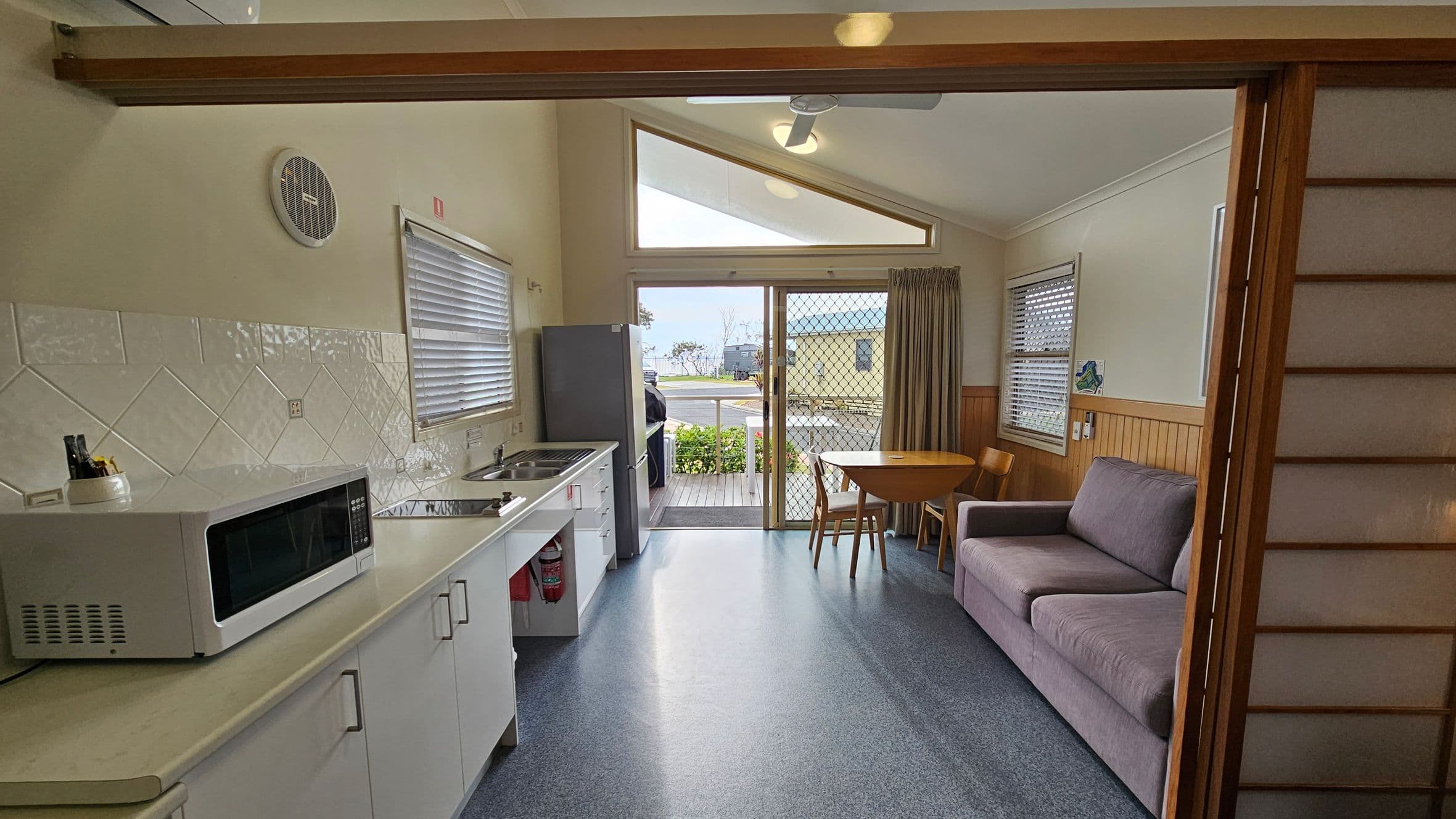 A compact kitchen and living area with a light-coloured countertop on the left, featuring a microwave and a sink. Above the counter, there are white tiles and a small round vent. A window with blinds is above the sink. In the centre, a sliding glass door with a mesh screen leads to an outdoor area. On the right, a small round table with chairs and a purple sofa are positioned against a wall with wood panelling. The ceiling is sloped with a fan and light fixture.