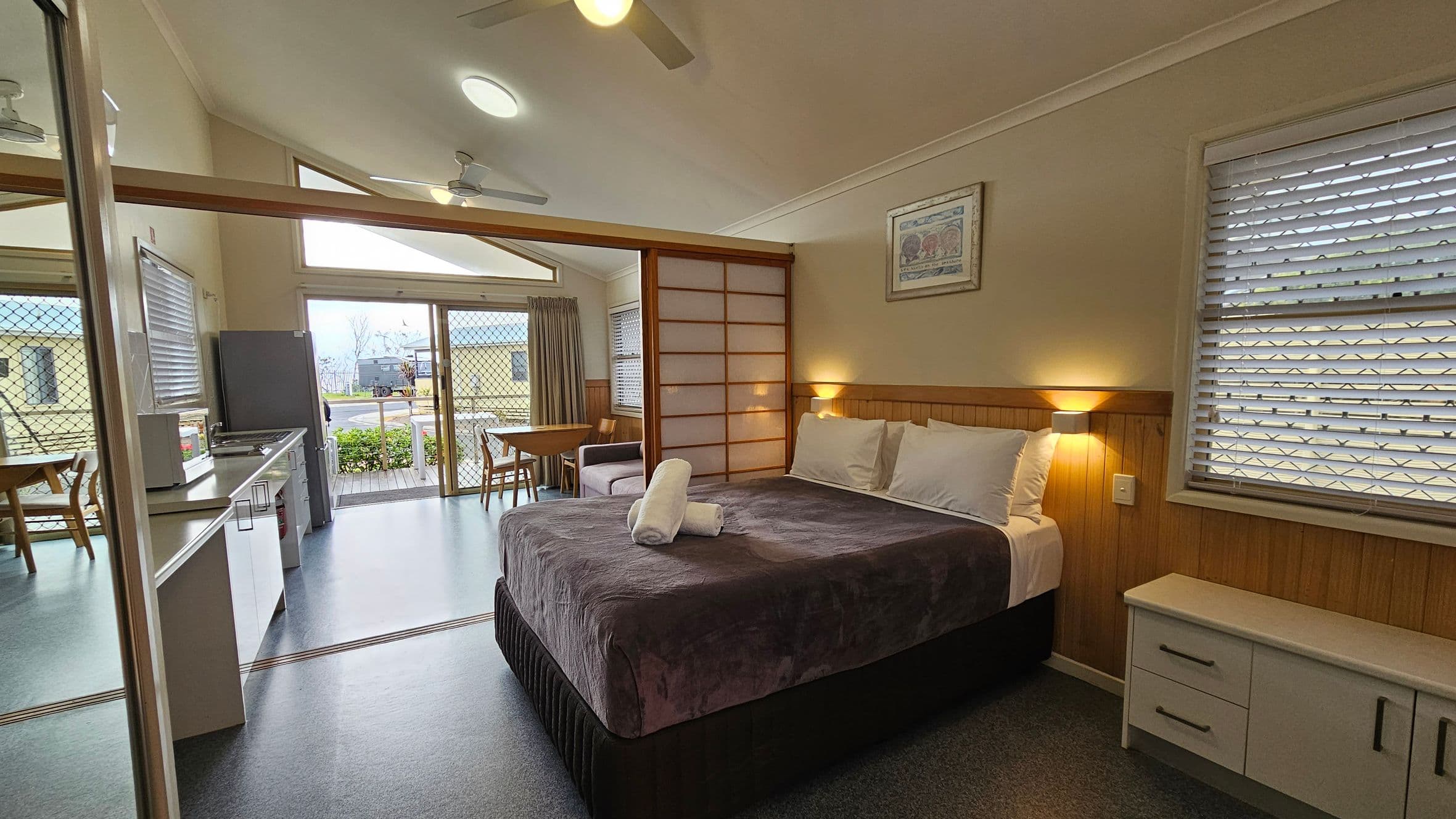 A room with a bed on the right, covered in a dark purple blanket with two rolled white towels on top. A small bedside table with drawers is next to the bed. On the left, there's a kitchenette with a sink, microwave, and a fridge. A dining table with chairs is near the sliding glass doors, which lead to an outdoor area. The ceiling is vaulted with a fan, and the floor is covered in a grey material. Windows with blinds are on the right wall.