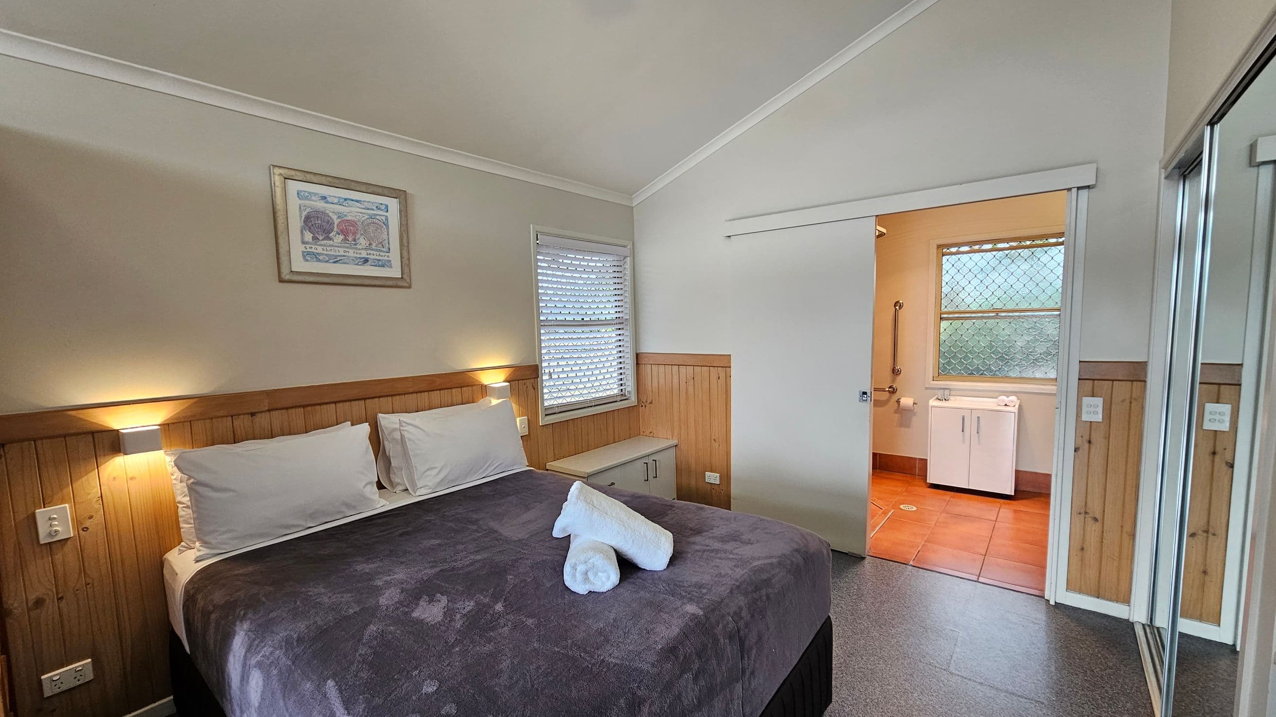 A bedroom with a double bed in the centre, covered with a dark blanket and two white towels on top. The headboard is wooden, with wall-mounted lights on either side. A framed picture hangs above the bed. To the right, a sliding door leads to a bathroom with a window and a white cabinet. A window with blinds is on the left wall. The floor is carpeted, and there is a mirrored wardrobe door on the right. The ceiling is sloped and painted white.