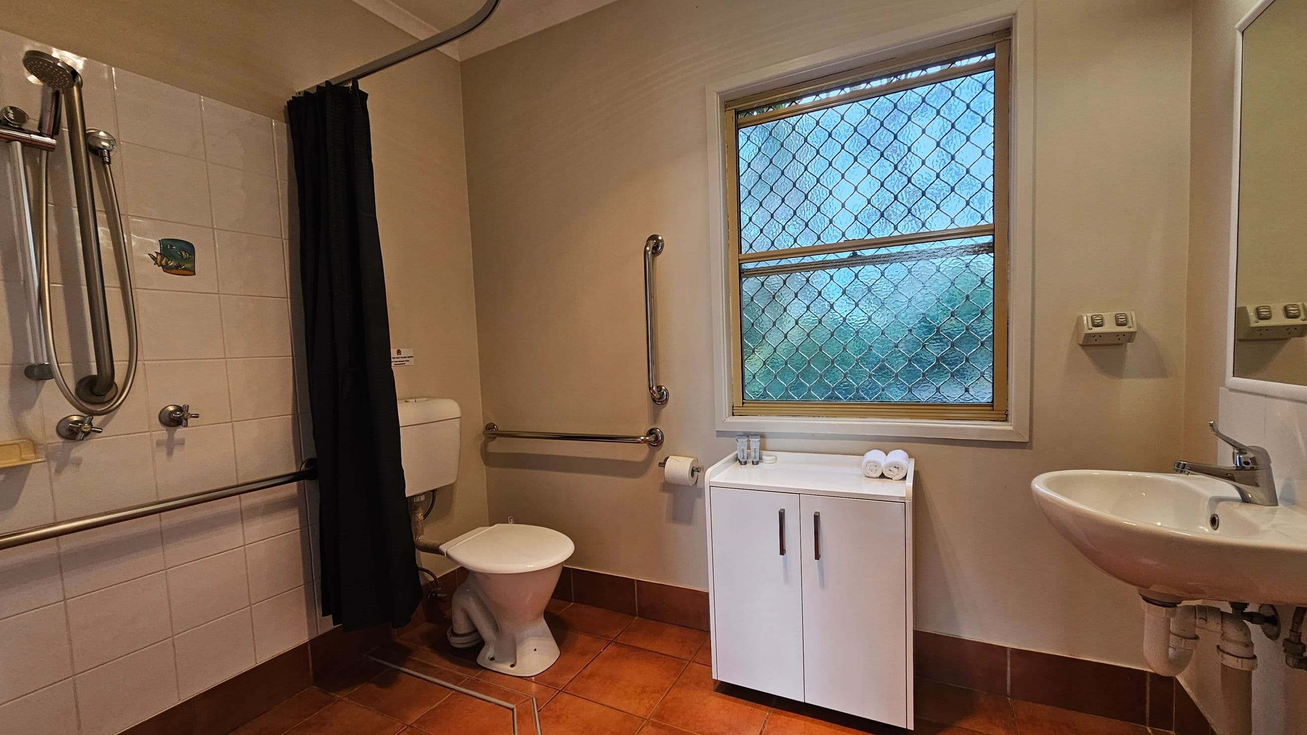 A bathroom with terracotta floor tiles and a white ceiling. On the left, a shower area with a handheld showerhead and a black curtain. A toilet with grab bars is next to it. In the middle, a window with a mesh screen above a white cabinet holding rolled towels and toiletries. On the right, a white sink with a mirror above it. The walls are light-coloured, and there is a small control panel on the wall near the sink.