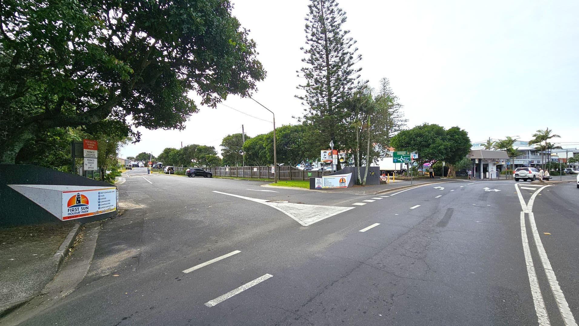 A road with a split, where the left side leads to a quieter street lined with trees and a sign for "First Sun Holiday Park." The right side is a busier road with multiple lanes and a few cars. There are signs and a small building on the right, with palm trees and a larger building in the background. The road surface is marked with arrows and lines, and the area is surrounded by greenery.