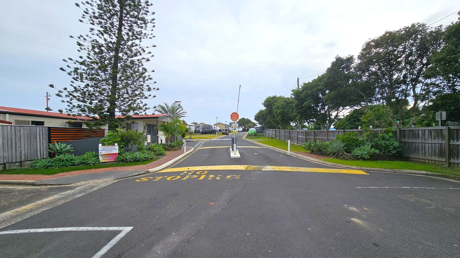 A road with a speed bump and "STOP" painted on it leads to a barrier gate. On the left, there's a tall tree and a building with a red roof, surrounded by a wooden fence and lush plants. A sign indicates reception. On the right, a wooden fence runs alongside the road, with more greenery and trees. In the background, there are more buildings and a green vehicle parked near the road.