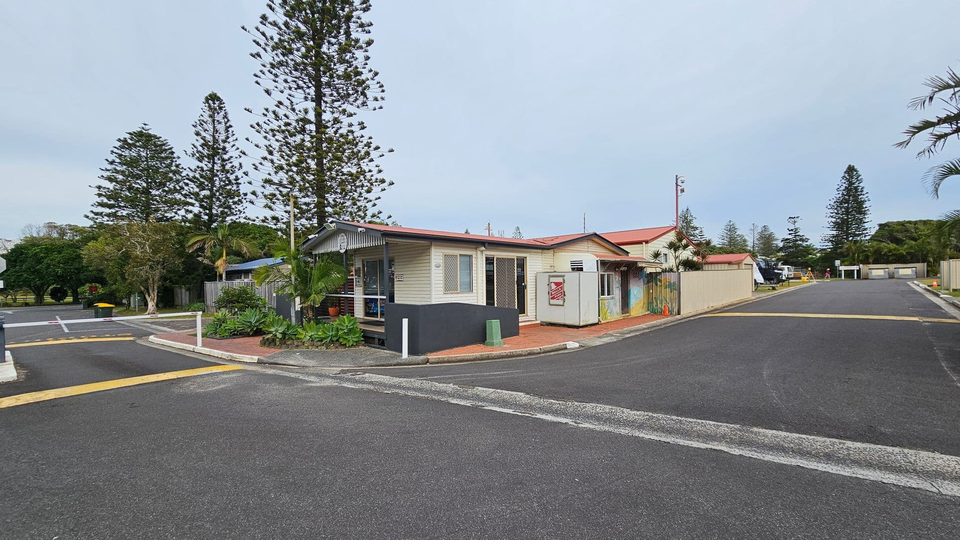 A small building with a red roof and light-coloured walls is situated at the corner of a paved road. The building has a porch with potted plants and a sign on the wall. Tall trees, including palm trees, are in the background. The road is wide and has a speed bump and a yellow bin on the left. The area is surrounded by greenery, with more buildings and a few vehicles visible further down the road. The sky is overcast.