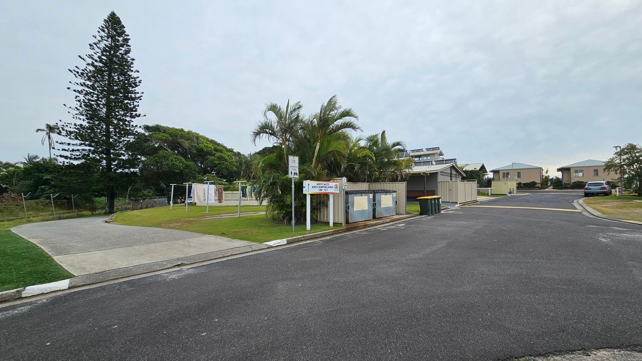 A residential area with a paved road in the foreground. On the left, there's a large coniferous tree and a grassy area with a clothesline. In the centre, palm trees and a sign are visible near a fenced area with bins. On the right, several houses with light-coloured walls and green roofs are seen, along with a parked car. The background features more greenery and additional residential buildings.