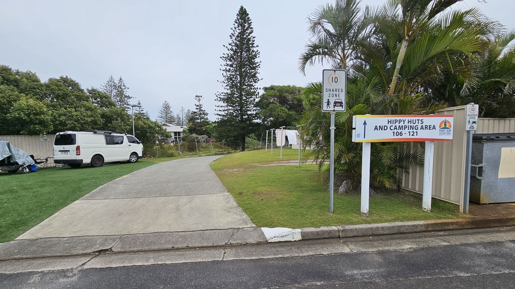 A paved path curves through a grassy area. On the left, a white van is parked on grass next to a tent covered with a tarp. Trees and a building are visible in the background. On the right, a sign reads "Hippy Huts and Camping Area 106-121" with a speed limit sign above it. Palm trees and a fence are nearby, with a bin next to the fence. The area is surrounded by greenery, including tall trees.
