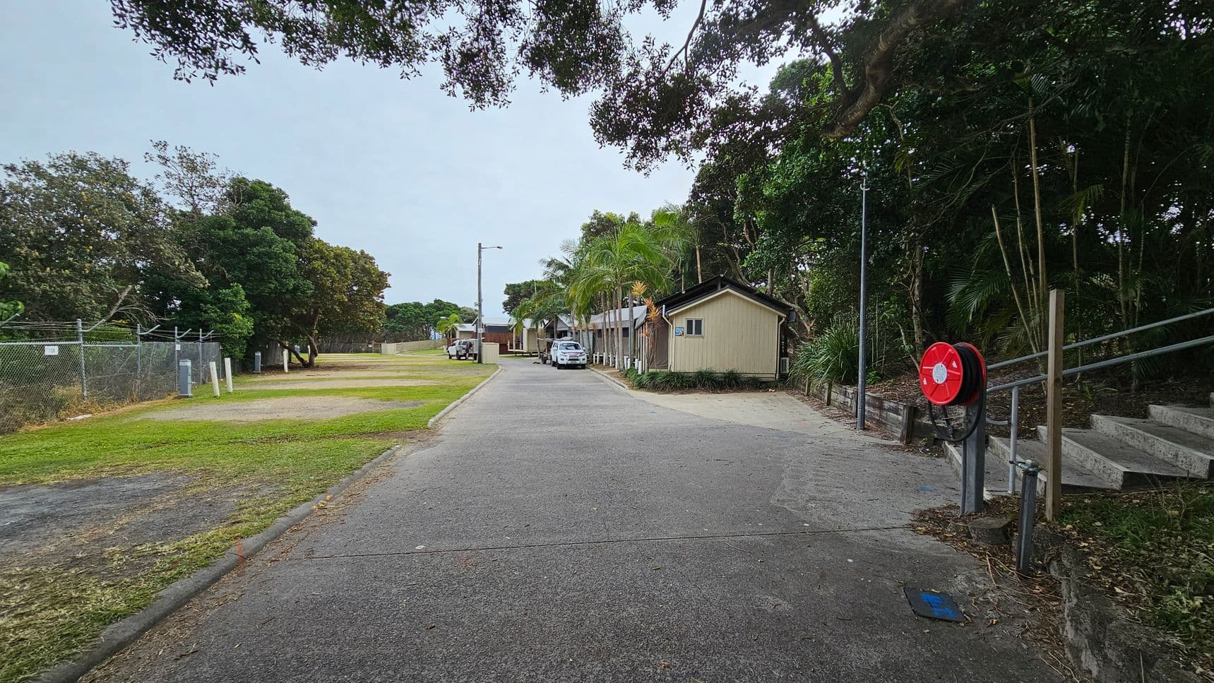 A paved path leads towards a group of small buildings surrounded by trees. On the right, a red fire hose reel is mounted near a set of steps. Palm trees and other greenery line the right side, while a car is parked further down the path. On the left, a grassy area is bordered by a chain-link fence with barbed wire. The area is shaded by overhanging tree branches.