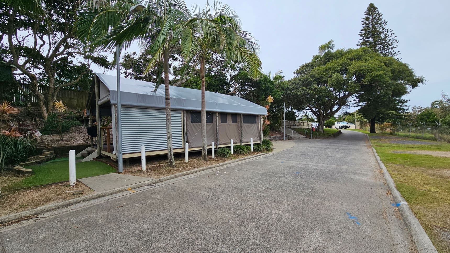 A small, corrugated metal building with a sloped roof is on the left, surrounded by palm trees and other vegetation. The building is elevated slightly above the ground with a ramp leading up to it. A paved road runs alongside the building, curving to the right. In the background, there are more trees, including a tall conifer, and a couple of parked vehicles. The ground is a mix of concrete and grass.