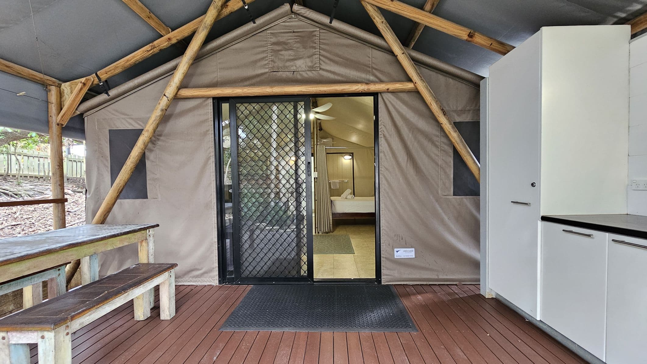 A rustic interior with a wooden floor and a ceiling supported by wooden beams. On the left, there's a wooden table with benches. In the centre, a sliding glass door with a mesh screen leads to a room with a visible bed and ceiling fan. On the right, there are white cabinets with black countertops. The walls are made of canvas, giving a tent-like appearance. A rubber mat is placed in front of the door.