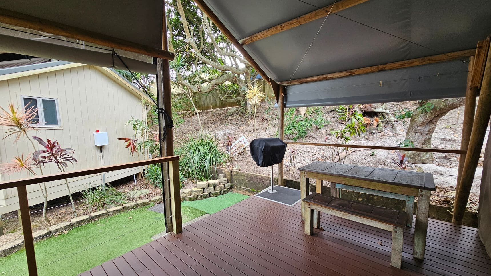 A covered outdoor area with a wooden deck and a picnic table with benches on the right. A barbecue grill with a cover stands nearby. The background features a sloped garden with various plants and a large tree. On the left, a light-coloured building with a small window is visible. The ground is a mix of artificial grass and natural soil, and the ceiling is made of a grey fabric supported by wooden beams.