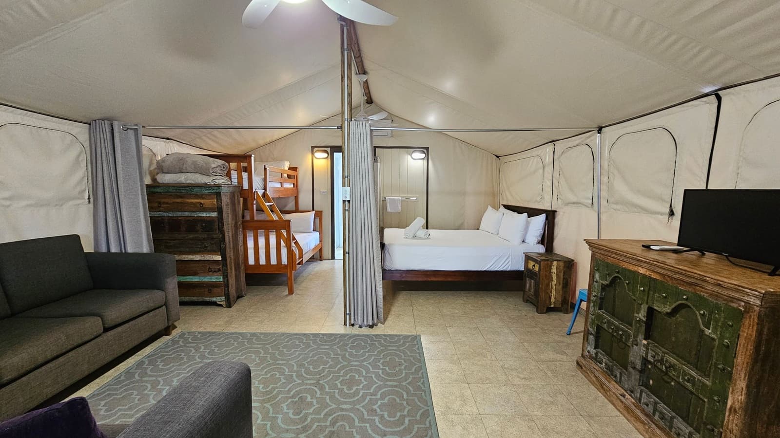 A spacious tent interior with a beige fabric ceiling and walls. On the left, there's a dark grey sofa and a patterned grey rug on a tiled floor. In the centre, a wooden bunk bed is visible, separated by a curtain from a double bed with white linens. On the right, a rustic wooden cabinet holds a flat-screen TV. The tent is well-lit with ceiling lights, and a fan is visible above.