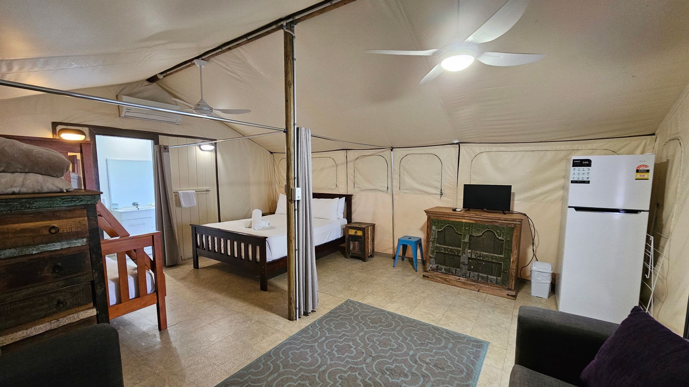 A spacious room with a beige tent-like ceiling and walls. On the left, there's a wooden bunk bed with folded towels on top. In the centre, a double bed with white linens is next to a small wooden nightstand. A curtain divides the space. On the right, a green rustic cabinet holds a small TV, with a blue stool beside it. A large white refrigerator stands nearby. The floor is tiled, and a patterned grey rug is in the foreground. Ceiling fans are mounted above.