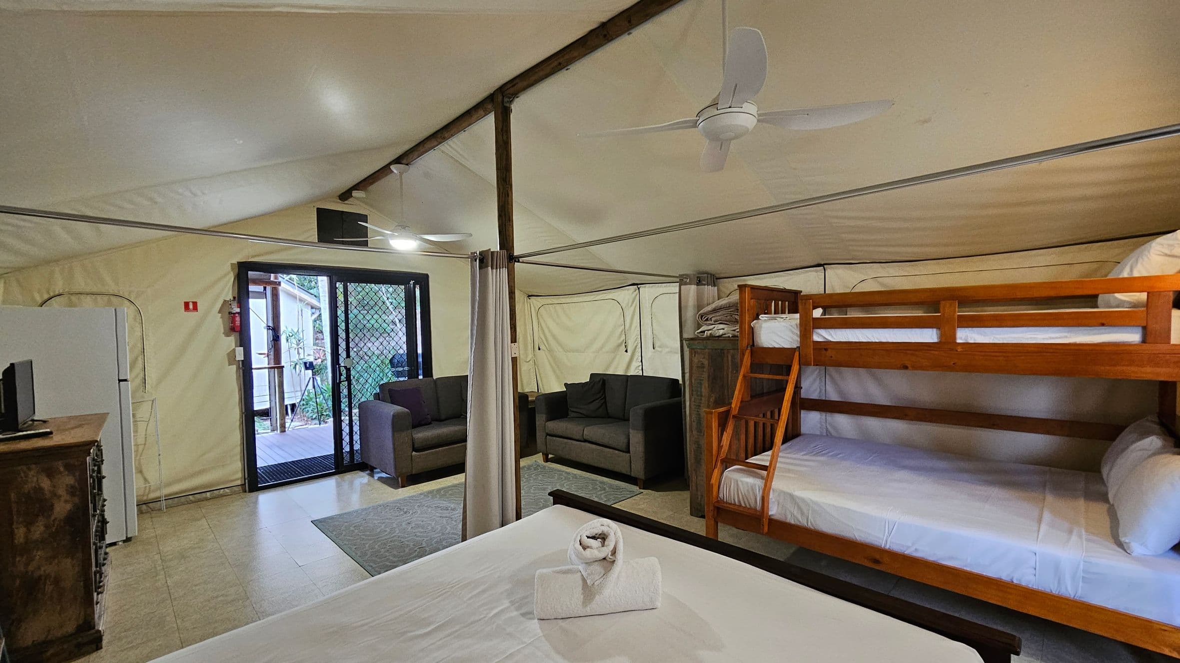 A spacious room with a high, sloped ceiling and a tiled floor. On the right, there is a wooden bunk bed with white bedding. In the centre, a bed with a rolled towel on top. To the left, a small refrigerator and a wooden cabinet with a TV. Near the entrance, two dark sofas are positioned by a sliding glass door with a mesh screen, leading outside. A ceiling fan is mounted above, and a curtain divides the space.