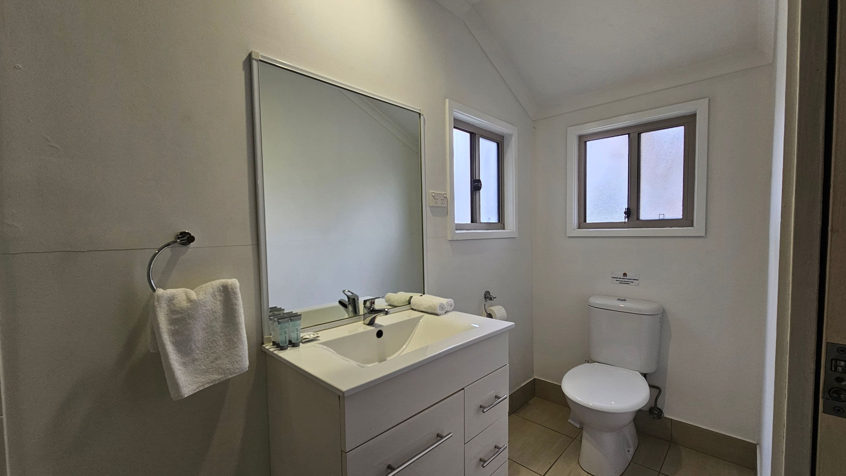 A small bathroom with a white vanity and sink on the left, featuring a mirror above and toiletries on the counter. A towel hangs on a ring to the left of the sink. In the centre, there are two small windows with white frames. On the right, a white toilet is positioned beneath the windows. The floor is tiled in a light brown colour, and the walls are painted a light shade.