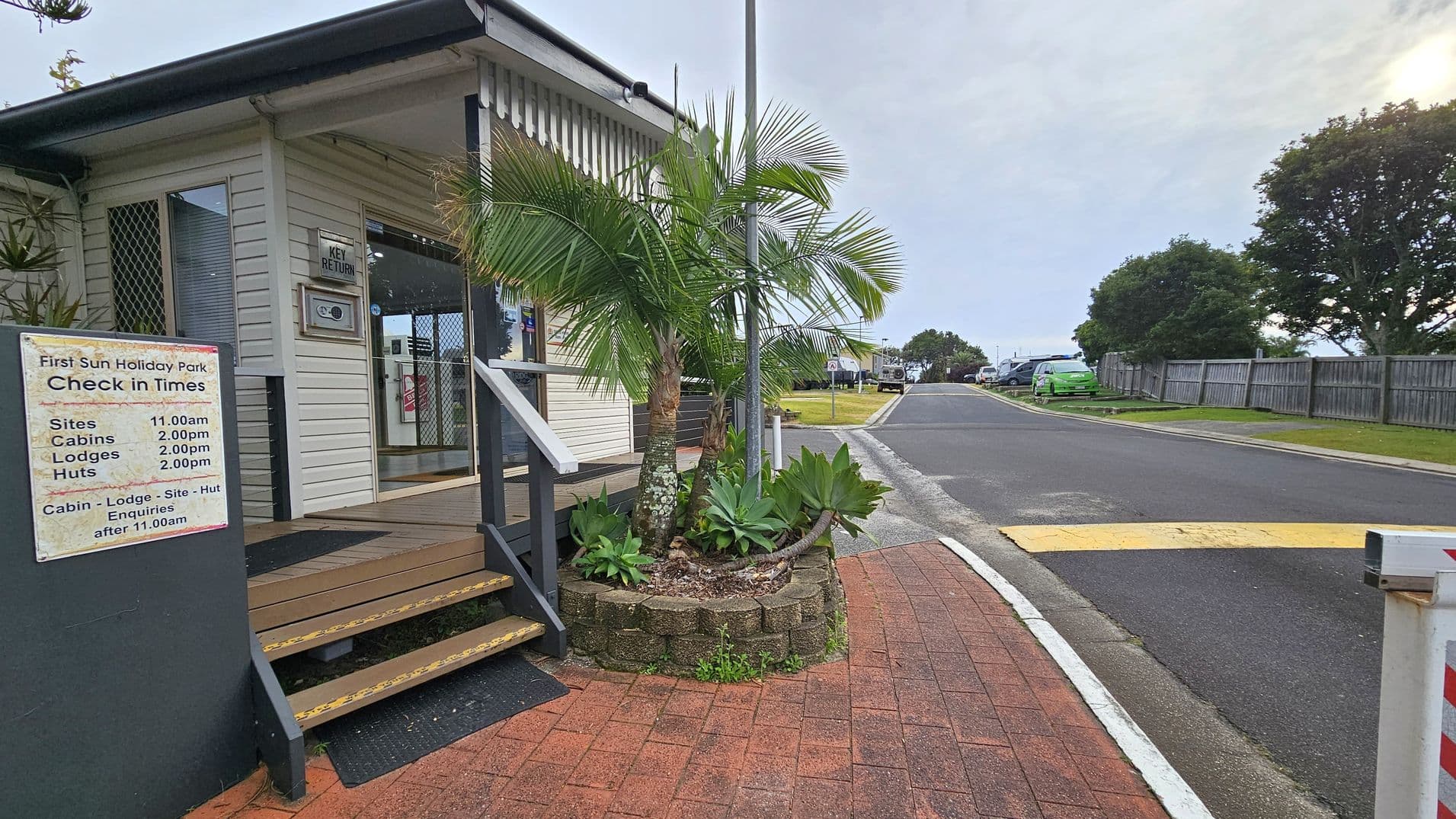 A holiday park entrance with a sign displaying check-in times is on the left. A small building with a porch and steps is adjacent to the sign. In front, a landscaped area features palm trees and succulents. A paved path leads to a road on the right, bordered by a wooden fence and grassy areas. Several vehicles are parked along the road, and trees are visible in the background.