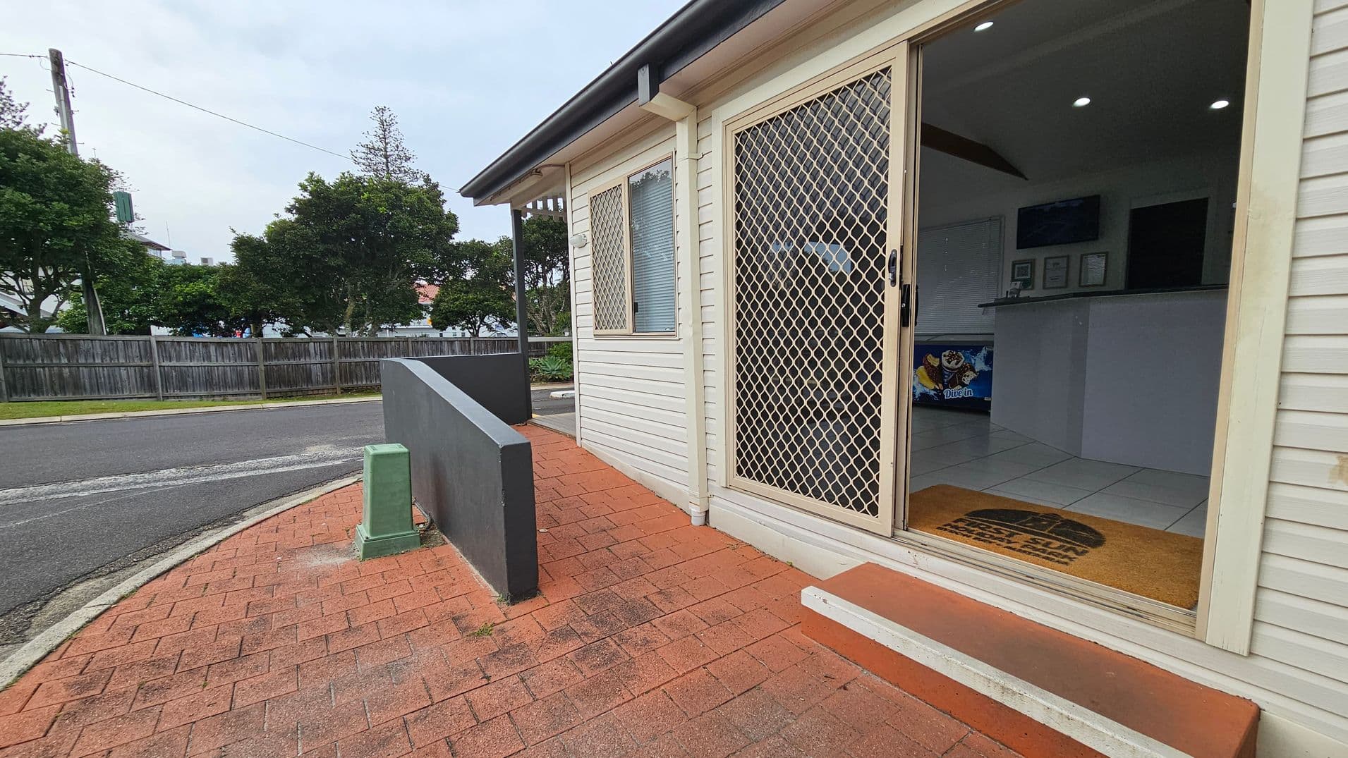A building with a white exterior and a sliding mesh door on the right. The door is open, revealing a tiled interior with a counter and some framed items on the wall. A doormat is placed at the entrance. The foreground has a red brick pathway leading to the door, with a small green utility box and a low grey wall. In the background, there is a street lined with trees and a wooden fence.