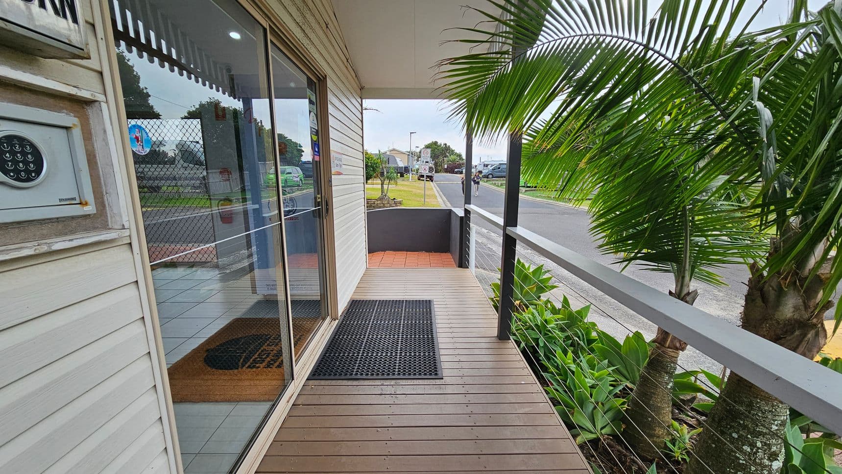 A wooden deck with a black mat leads to a glass sliding door on the left, reflecting the street and nearby greenery. A keypad is mounted on the wall beside the door. On the right, lush palm trees and plants line the edge of the deck, separated by a white railing. The street in the background has a few parked cars and a cyclist. The ceiling is plain and white, and the floor is wooden with a tiled section near the entrance.