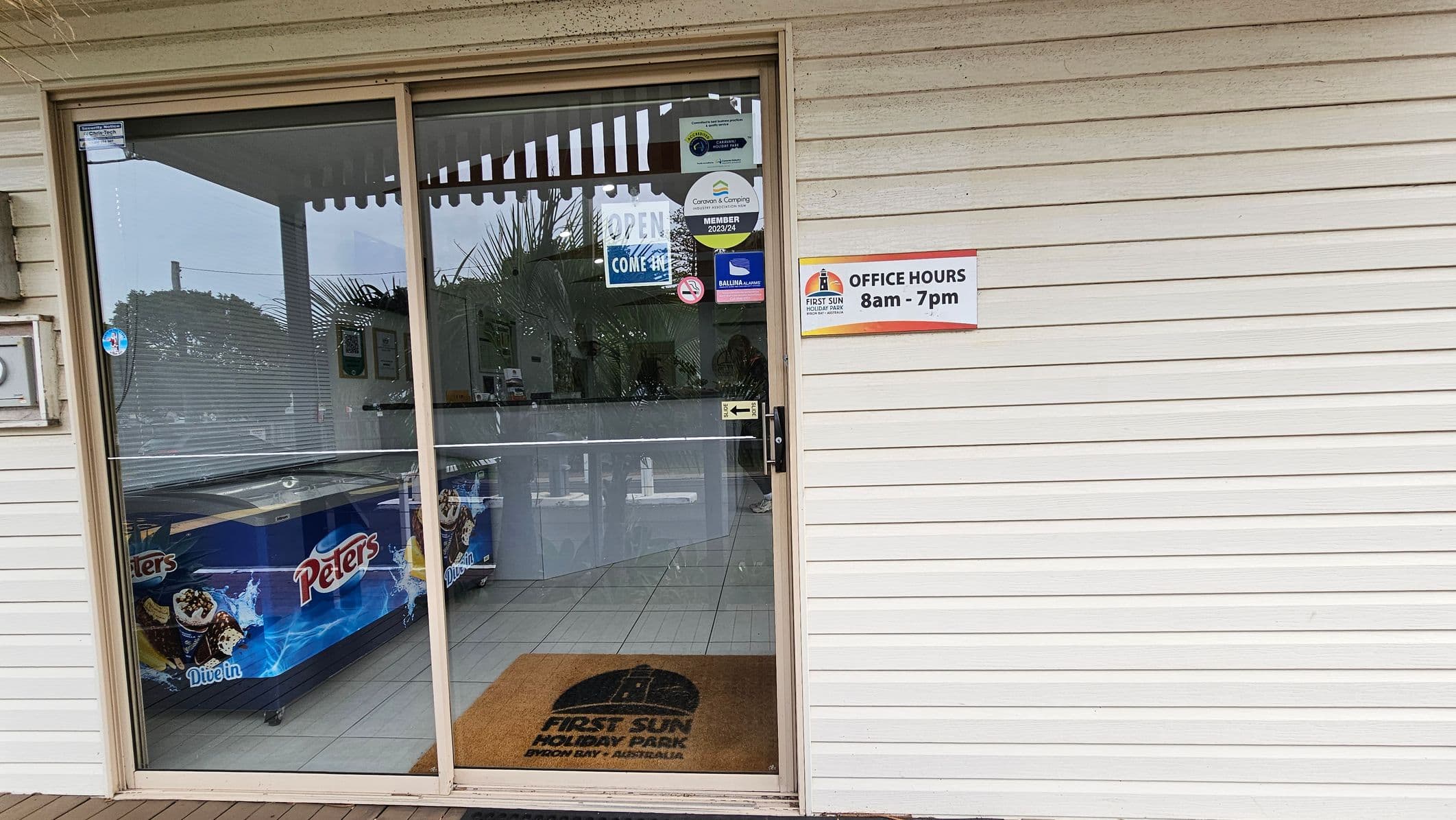 A glass door with various stickers, including "OPEN COME IN" and membership signs, is on the left. Behind the door, there's a freezer with "Peters" branding. A doormat with "FIRST SUN HOLIDAY PARK" is on the floor. On the right, a white wall features a sign indicating office hours from 8am to 7pm. The floor is tiled, and the ceiling is not visible.