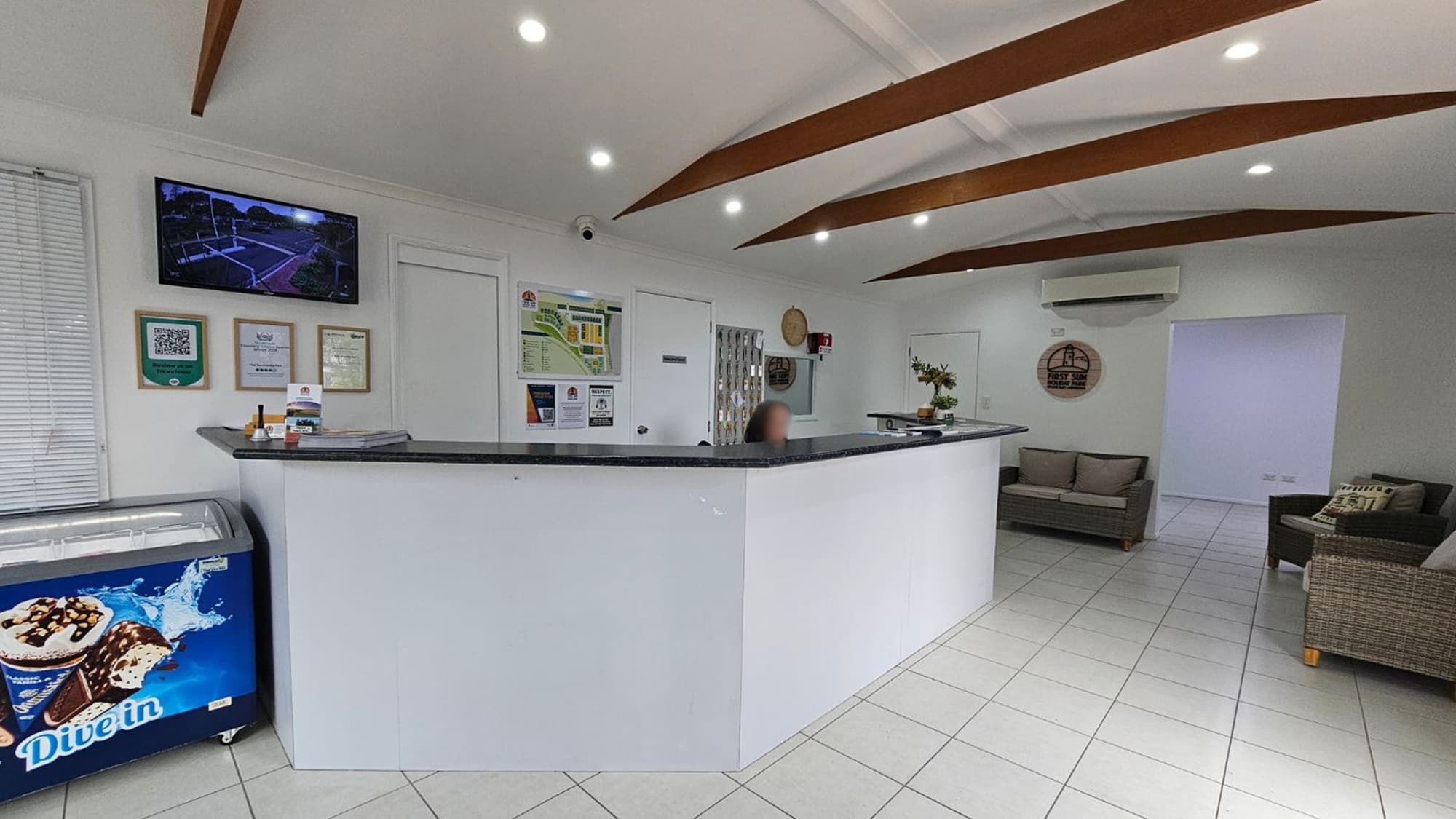 A reception area with a white, L-shaped counter in the centre. On the left, there's an ice cream freezer with a "Dive in" graphic. Above it, a TV is mounted on the wall, alongside several certificates and a QR code. The right side features a seating area with grey sofas and a wicker chair. The ceiling has wooden beams and recessed lighting, while the floor is tiled in white. An air conditioning unit is mounted on the wall above the sofas.