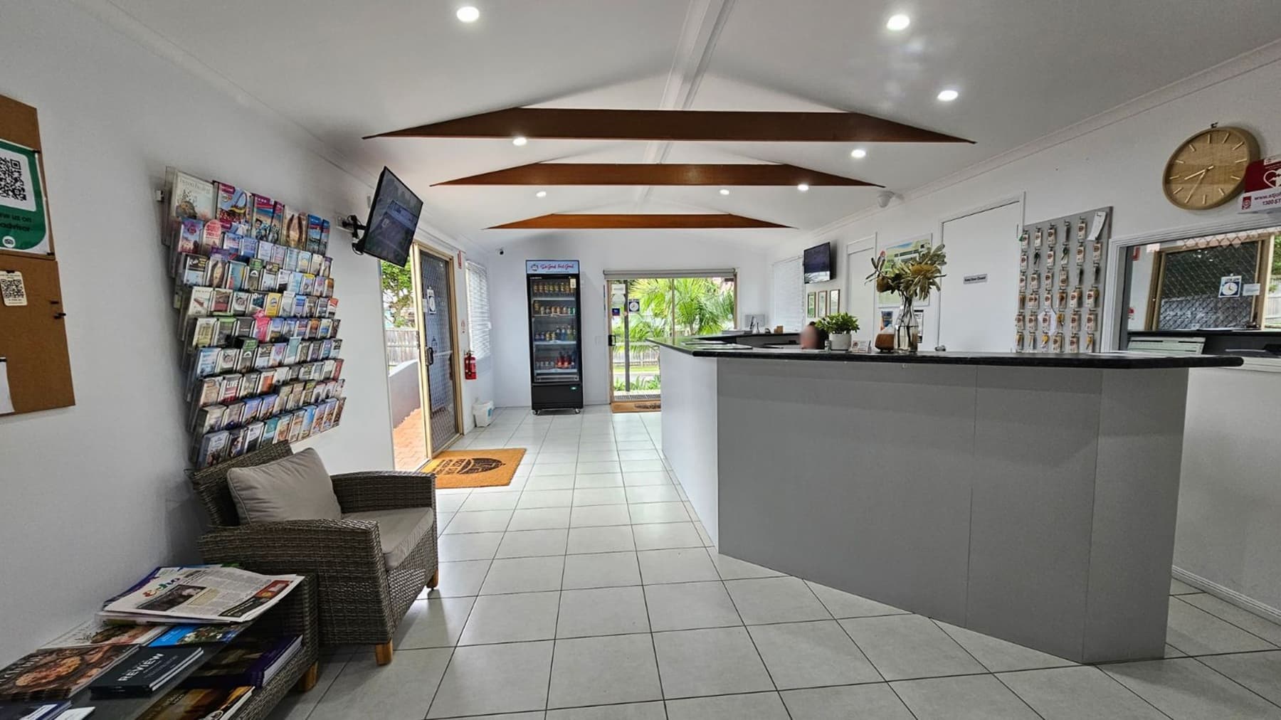 A reception area with a white tiled floor and a ceiling featuring wooden beams. On the left, there's a wall-mounted display of brochures and a grey armchair with a small table holding magazines. A television is mounted above. In the centre, a glass door leads outside, flanked by a vending machine. On the right, a curved reception desk with plants and a clock on the wall behind it. The space is well-lit with recessed lighting.