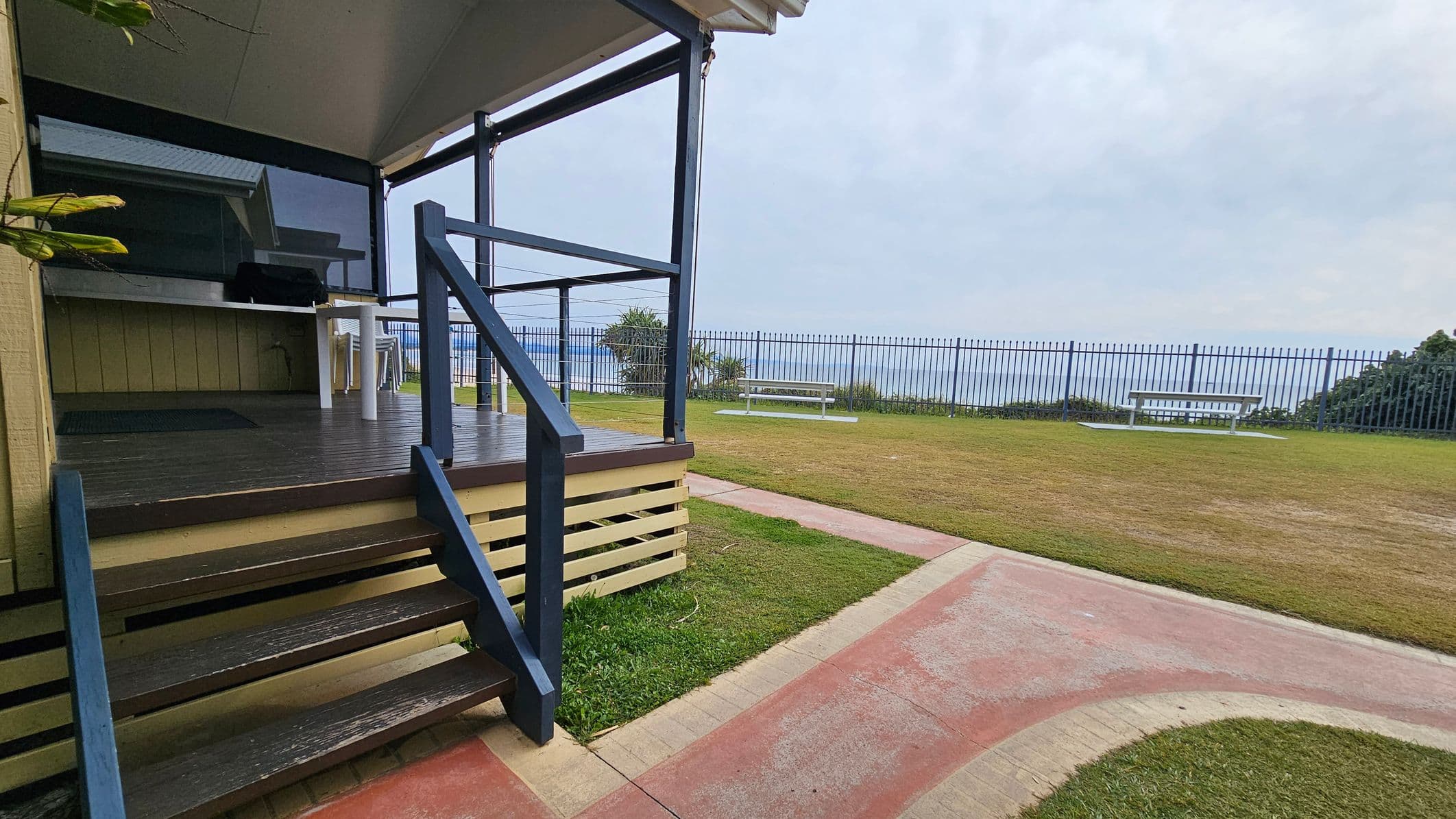 A wooden deck with dark steps and a railing is on the left, leading to a covered area with a table and chairs. The ground is a mix of grass and a paved path. In the background, a metal fence runs along the edge of a grassy area, with a white bench facing the sea. The sky is overcast.