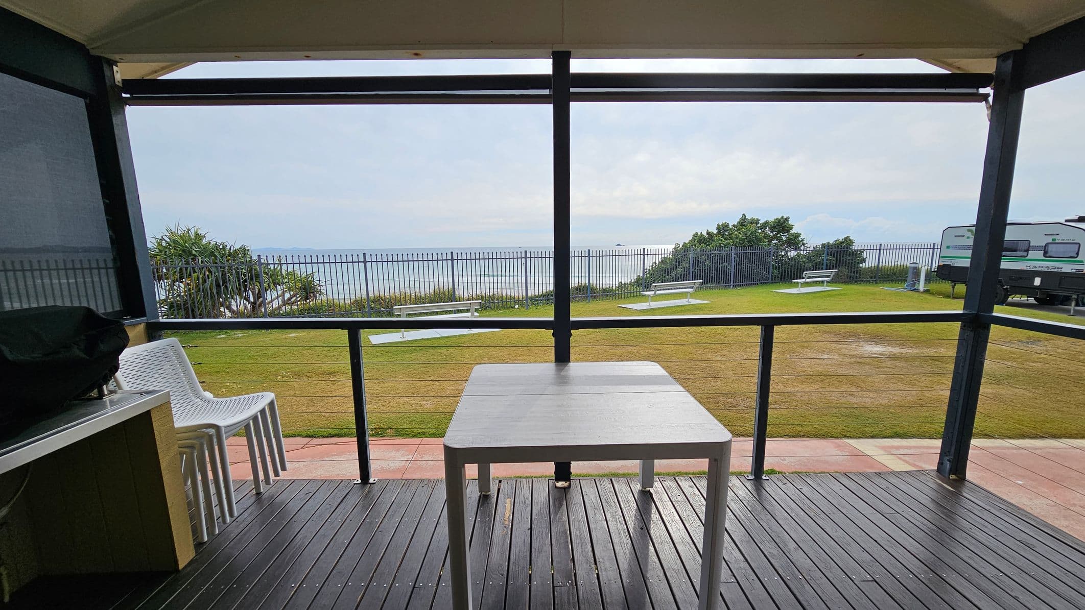 A covered outdoor area with a dark wooden floor and a white table in the centre. On the left, there are stacked white chairs. Beyond the railing, a grassy area with white benches is visible. In the background, a metal fence runs along the edge, with a view of the sea and distant hills. On the right, a caravan is parked on the grass. The ceiling is light-coloured and the overall setting suggests a coastal location.