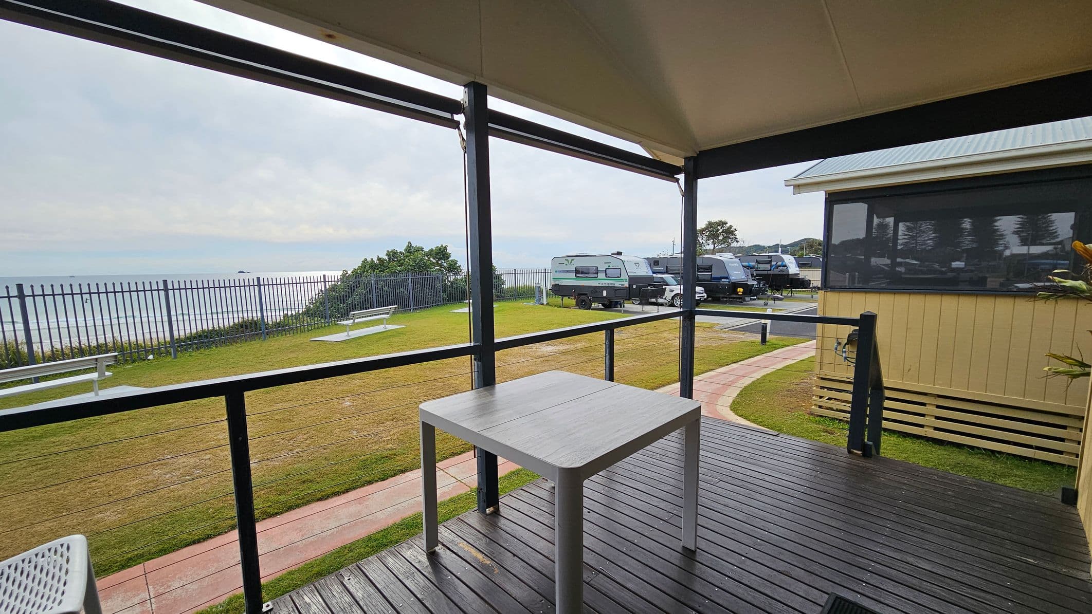 A covered deck with a wooden floor features a simple square table. Beyond the railing, a grassy area with benches overlooks the ocean, separated by a metal fence. On the right, a light-coloured building with large windows is visible. Several caravans are parked on a paved area in the background. The scene suggests a coastal setting with a mix of natural and recreational elements.