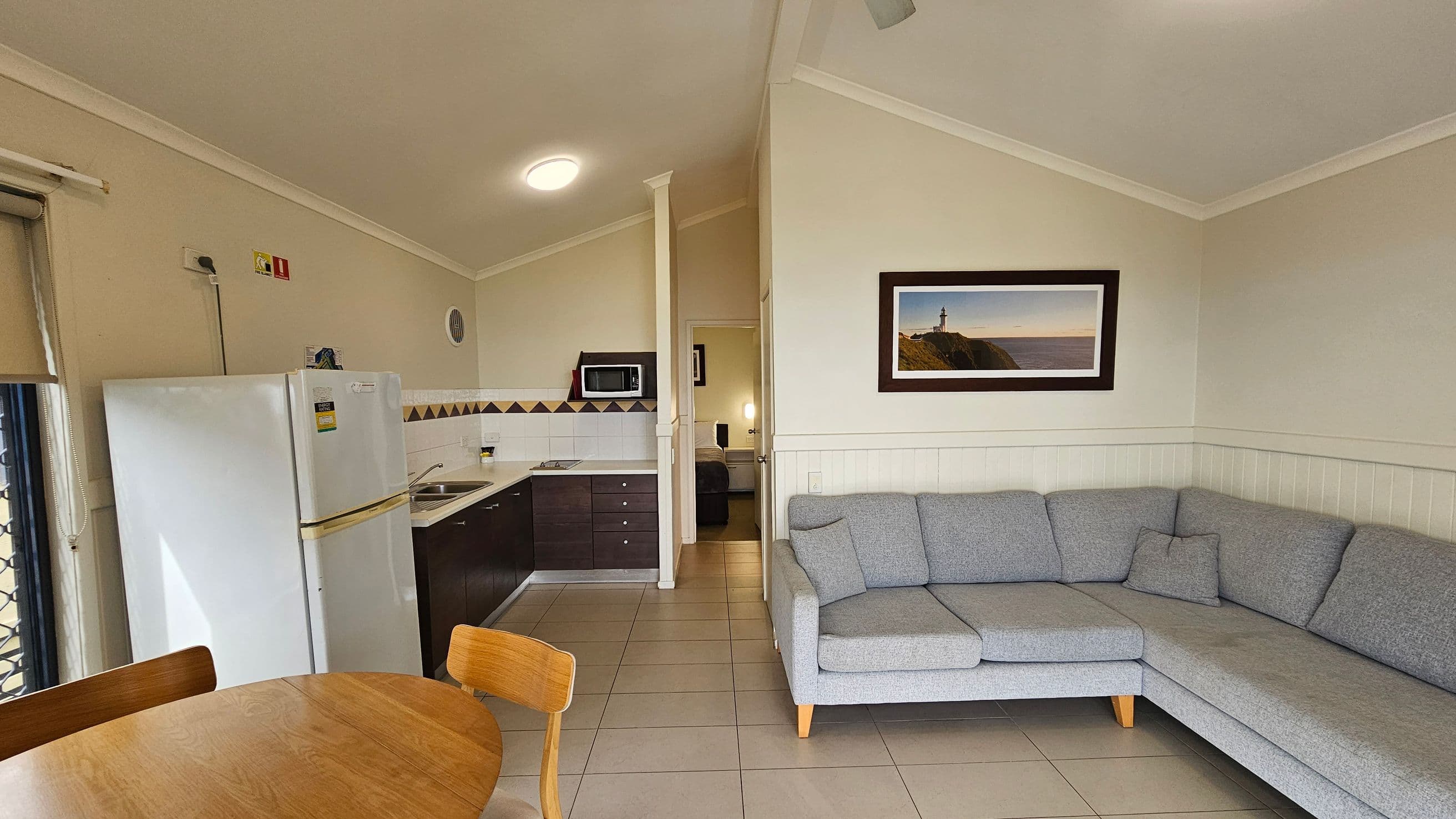 A small living area with a grey L-shaped sofa on the right, positioned against a cream wall with a framed picture of a lighthouse. In the centre, a doorway leads to another room. On the left, a kitchen area features a white fridge, a countertop with a sink, and a microwave. A round wooden dining table with chairs is in the foreground. The floor is tiled, and the ceiling is sloped with a ceiling fan.