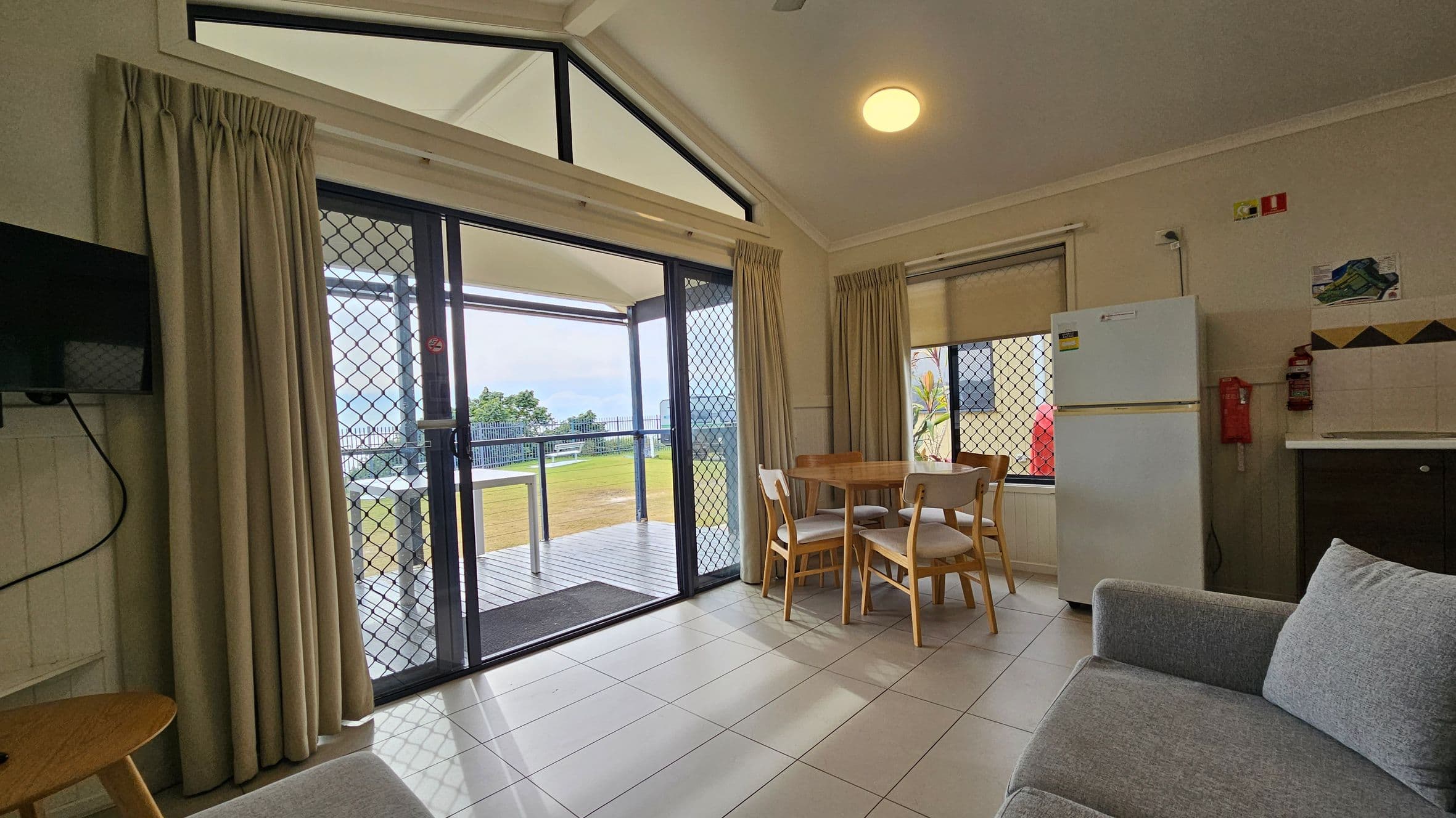 A room with a tiled floor and a high ceiling. On the left, a small TV is mounted on the wall above a round wooden table. In the centre, large glass doors with mesh screens open to a patio with a view of grass and trees. Beige curtains frame the doors. On the right, a wooden dining table with four chairs is near a window. A white fridge stands beside a small kitchen counter. A grey sofa is partially visible in the foreground.