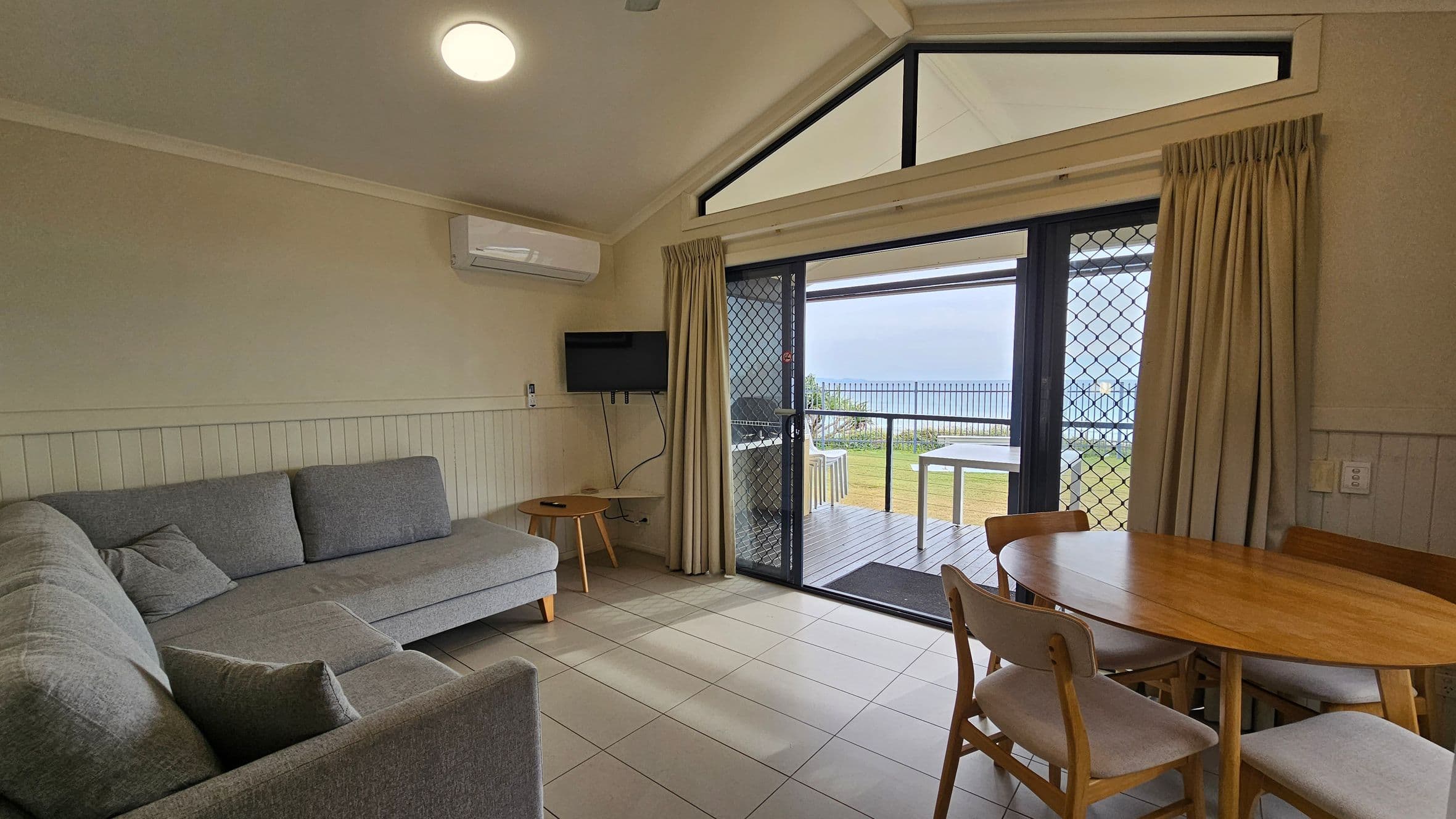 A cosy living room with a grey L-shaped sofa on the left, accompanied by a small round wooden side table. A wall-mounted TV is positioned above the table. On the right, a wooden dining table with four chairs is set near sliding glass doors leading to a patio. The doors have a mesh screen and are flanked by beige curtains. Outside, a grassy area and a railing are visible. The ceiling is vaulted with a triangular window above the doors, and the floor is tiled.