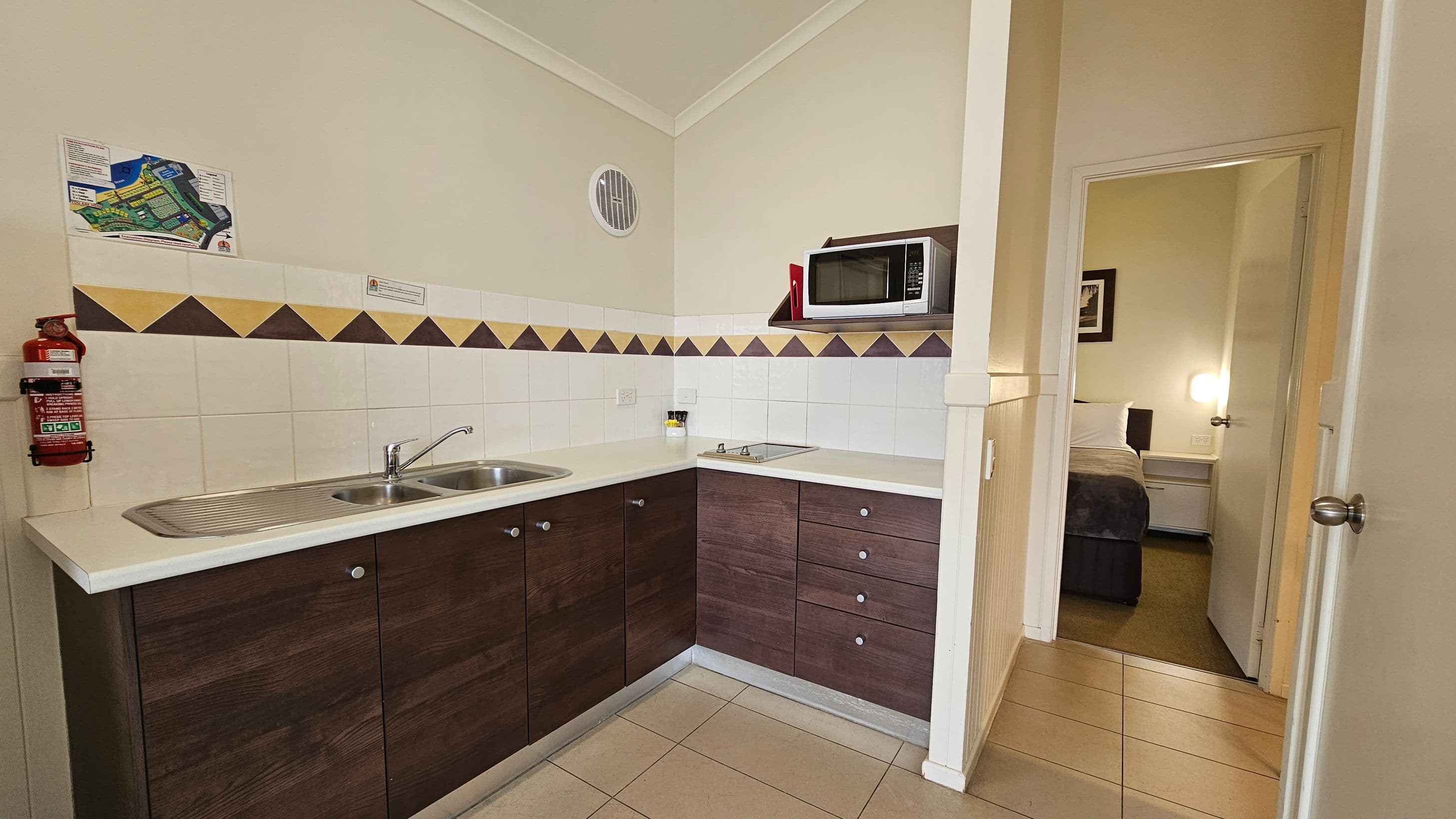 A compact kitchen area with a white countertop and dark wood cabinets. On the left, a double sink is installed, with a fire extinguisher mounted on the wall above. The backsplash features a pattern of brown and beige tiles. On the right, a microwave is placed on a shelf above a small stovetop. A doorway leads to a bedroom with a bed and a lit bedside lamp. The floor is tiled, and the ceiling is plain white.