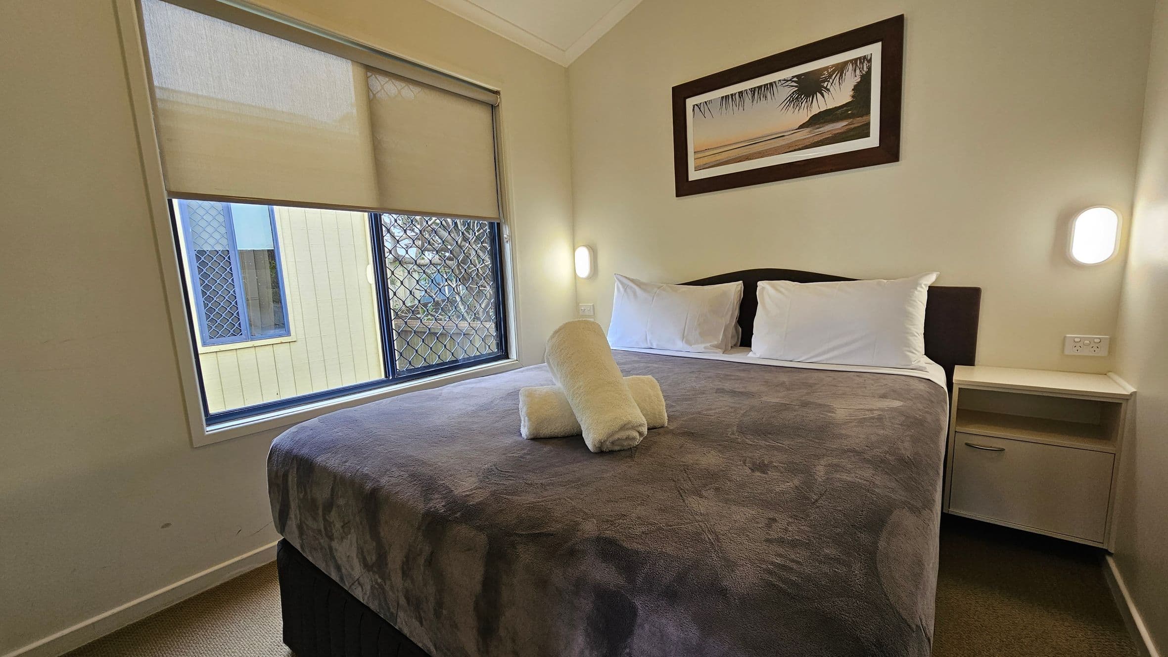 A small bedroom with a double bed in the centre, covered with a dark patterned blanket and two white pillows. Two rolled white towels are placed on the bed. On the right, a small bedside table with a drawer is beneath a wall-mounted light. Above the bed, a framed picture of a beach scene hangs on the wall. On the left, a window with a partially drawn blind and a security screen offers a view of a neighbouring building. The floor is carpeted in a neutral tone.