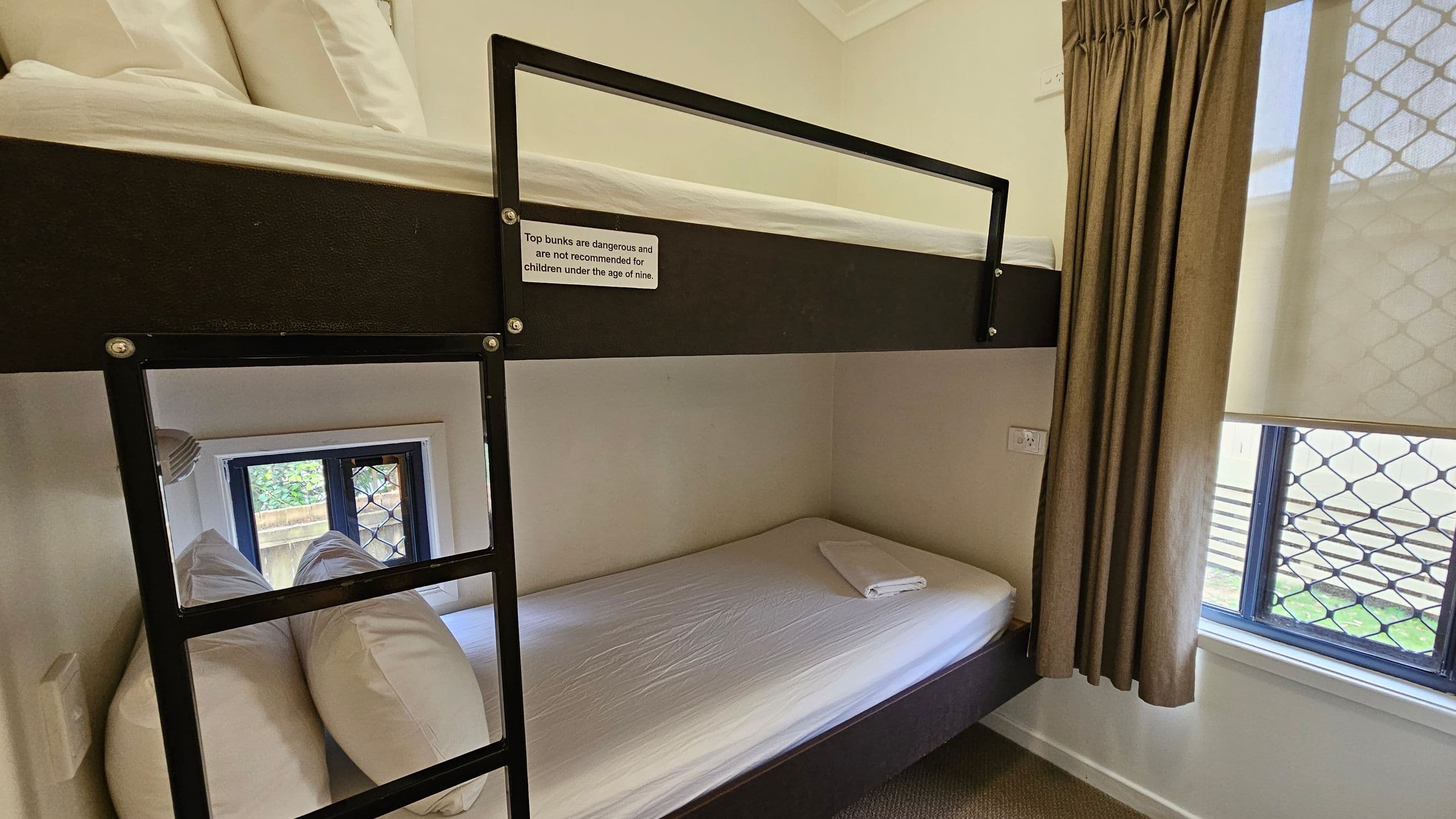 A room with a bunk bed against the wall. The top bunk has a safety rail and a sign warning against use by children under nine. The lower bunk is neatly made with a folded towel on the mattress. A ladder is attached to the bed frame. To the right, a window with a security grille is partially covered by a beige curtain. The floor is carpeted, and the walls are light-coloured.