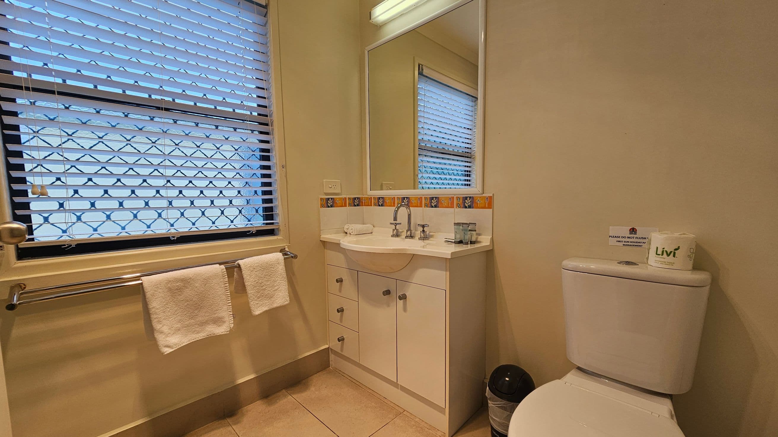 A bathroom with a window on the left featuring horizontal blinds and a security grille. Below the window, a towel rail holds two white towels. In the centre, a white vanity unit with a sink, chrome taps, and a mirror above. The backsplash has a decorative tile border. On the right, a white toilet with a small bin beside it. The floor is tiled in a light colour, and the walls are painted a soft beige.