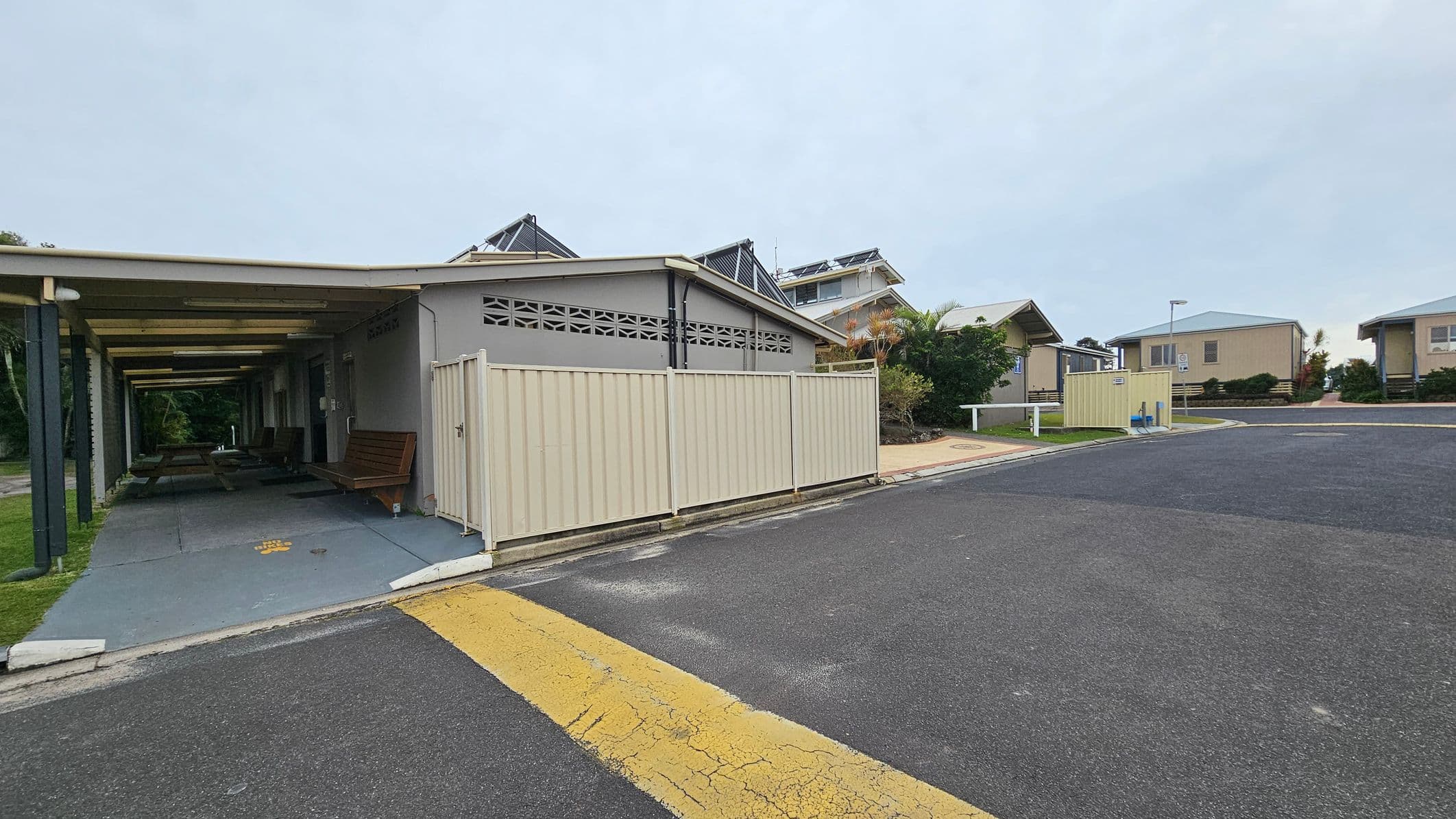 On the left, there is a covered walkway with a green roof supported by green pillars, leading to a building with a bench outside. The building has a grey wall with decorative ventilation blocks near the roof. In the centre, a light-coloured fence encloses part of the building. On the right, there is a paved road with a yellow speed bump, leading to several small houses with light-coloured walls and grey roofs. Bushes and small trees are scattered around the area.