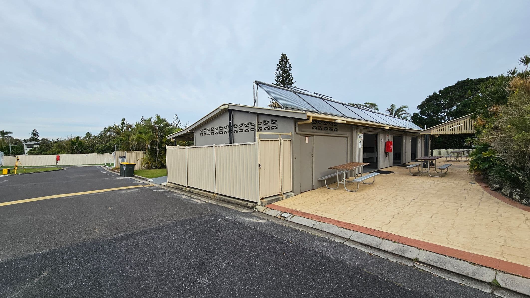 A building with a sloped roof featuring solar panels is on the right. It has a light-coloured fence and a small gate. In front, there are picnic tables on a paved area. A red fire extinguisher is mounted on the building's wall. On the left, a paved road leads to a fenced area with greenery, including palm trees. The background has more trees and a tall conifer. The ground is a mix of asphalt and light-coloured paving.